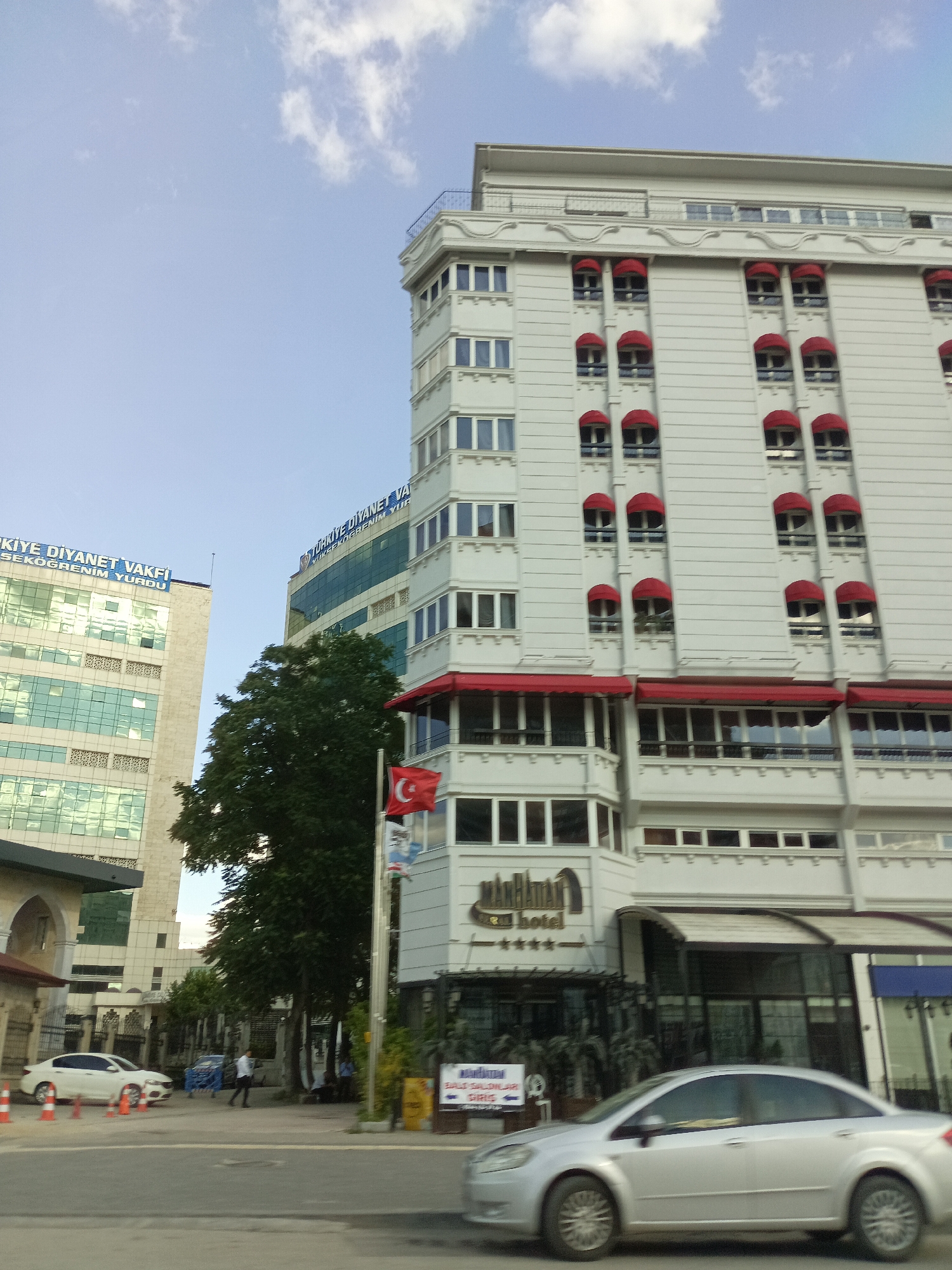 The image shows a street scene in Altındağ, Turkey, dominated by modern multi-story buildings. A white sedan car is in the foreground, moving from left to right. To the left of the sedan, an orange traffic cone is visible on the road. Behind the car, another modern building with a glass facade and the text "TÜRKİYE DİYANET VAKFI İSEKOĞRENİM YURDU" is visible. Further to the right, a taller, white building with numerous red awnings over its windows stands prominently. This building has the name "Manhattan Hotel" visible on its facade, along with a Turkish flag on a pole. Green trees are present between the buildings, and the sky is partly cloudy. In the background, another building with a glass facade is visible, and a person in a white shirt and dark pants is walking on the sidewalk. Several other traffic cones are scattered on the side of the road.