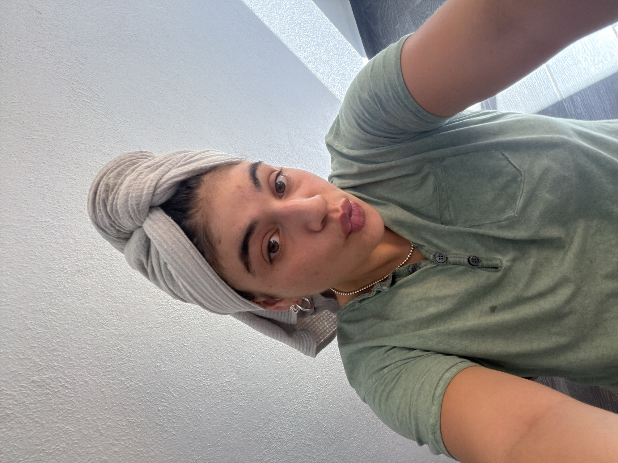 A medium close-up, diagonal selfie captures a young woman indoors. She has dark hair, wrapped in a light grey, ribbed towel, possibly after a shower. Her face is turned slightly towards the camera, with a playful "duck face" or puckered lips expression. Her brown eyes look directly into the lens. She wears a casual, faded olive-green Henley-style t-shirt with three black buttons on the placket and a single chest pocket on the left side. A silver beaded necklace is visible around her neck, and she has a silver hoop earring in her left ear.
The background is simple and mostly consists of a light-colored, textured wall, likely painted white or off-white. In the upper right corner, parts of a darker floor or wall and some natural light suggesting a window are visible, indicating it's daytime. The overall impression is casual and personal, likely taken in a home setting in Puebla, Mexico. There is no visible text in the image.