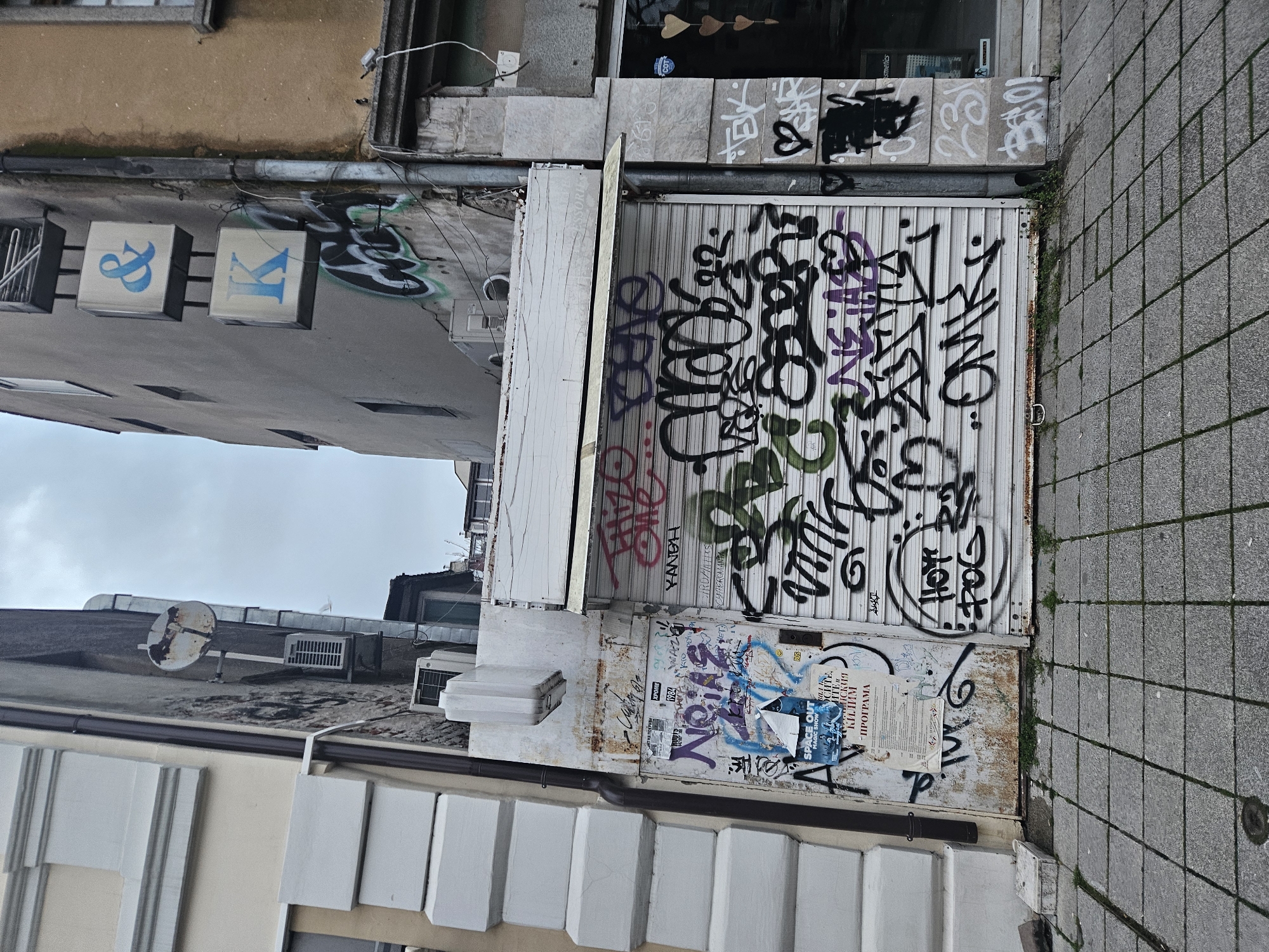 The image shows a narrow street scene in what appears to be an urban environment, likely in Burgas, Bulgaria, given the text on the posters and signs. The photograph is rotated, making it seem like the viewer is looking up at the side of a building. The primary subject is a storefront with a corrugated metal roll-down door covered in graffiti, featuring tags in black, green, and purple. A partially torn poster is affixed to the lower part of the door, containing text in Bulgarian and English ("SPACE OUT"). The adjacent wall is stained and covered with additional graffiti. The sidewalk is paved with grey square blocks. Above the storefront, there are building signs with "&" and "K" in blue. A satellite dish is seen on the roof of the building.