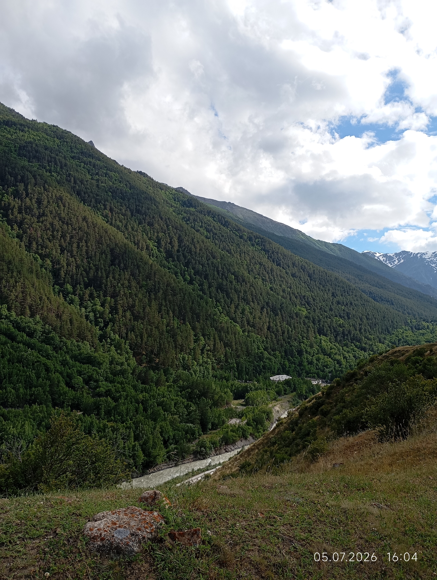 The image depicts a mountainous landscape, likely in the vicinity of Neutrino, Russia, as indicated by the timestamp. The foreground is dominated by a grassy slope with a few rocks, one of which is covered in lichen. Beyond the slope, a river winds through a valley. The valley walls are densely covered with evergreen trees, creating a dark green carpet that stretches up to the mountain peaks. In the distance, snow-capped mountains are visible against a sky filled with a mix of white and gray clouds. The overall scene is a natural panorama of a mountainous region. There are no people or animals visible in the image, and no human activity other than a few distant buildings barely visible through the trees in the valley.