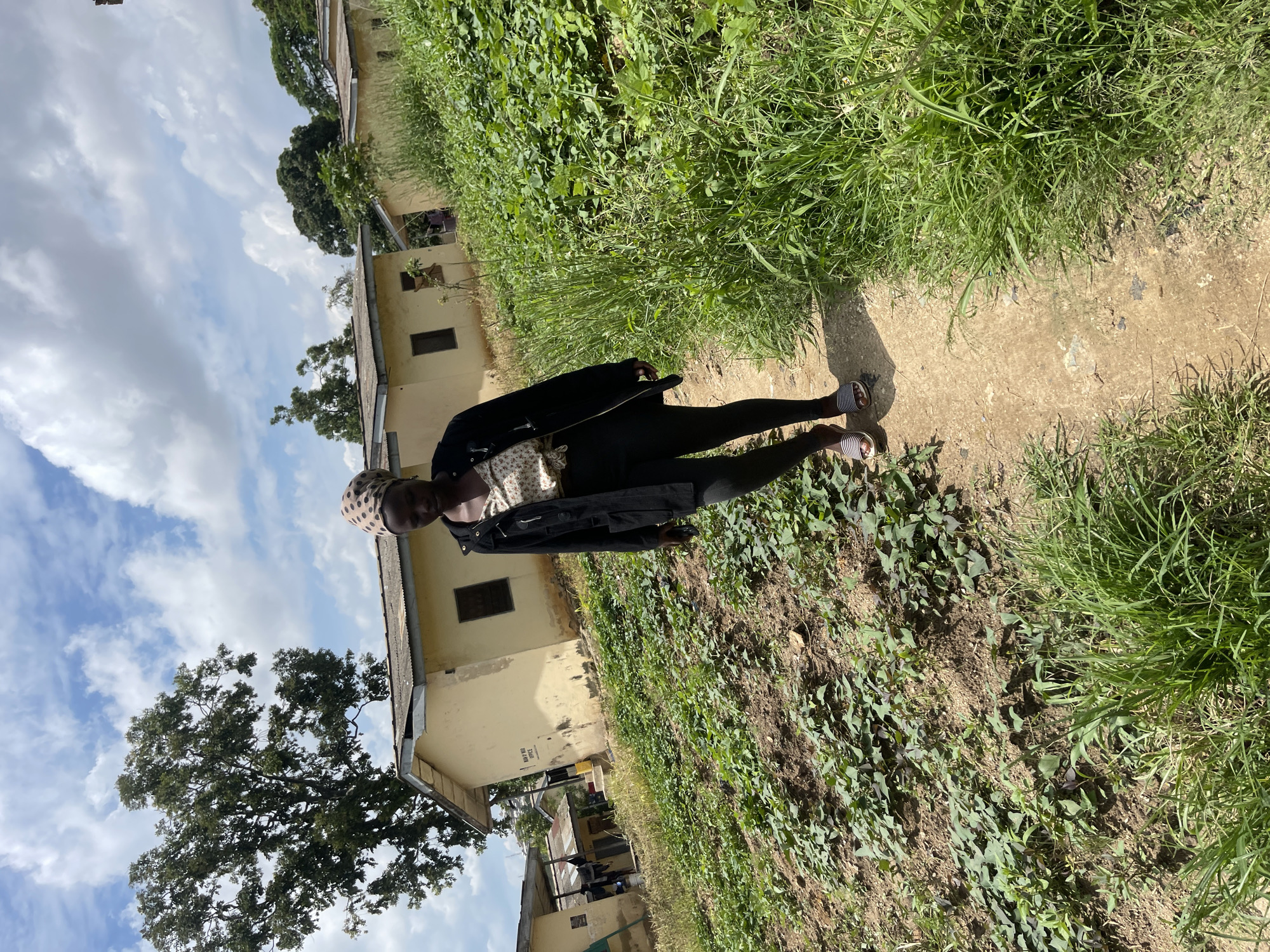 A young woman walks on a dirt path through an overgrown area with buildings in the background. She is wearing a black jacket over a patterned shirt and black leggings, with striped sandals on her feet. Her head is covered with a patterned scarf. To her left, a field of green plants is visible, and to her right, tall grass grows alongside the path. In the background, several single-story, light-colored buildings with corrugated metal roofs are situated on a slight incline. A large tree with dense foliage stands to the left of the buildings. The sky is partly cloudy with white cumulus clouds against a blue background. The scene appears to be a rural or semi-rural setting in the city of Rafin Par, Nigeria.
