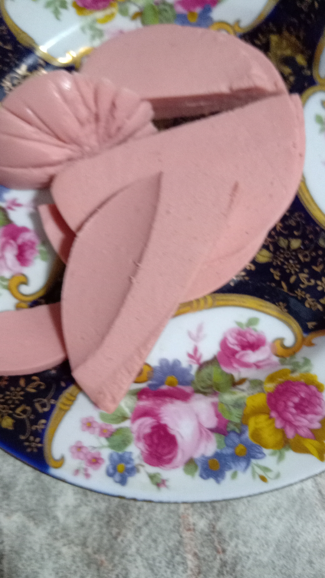 A sliced sausage arrangement rests on a floral-patterned plate. The plate features pink roses, blue flowers, and yellow accents against a white background. The plate also has a navy-blue border and gold trim. The plate rests on a grey textured surface. The scene is likely indoors. It is not possible to determine the location in Yerevan, Armenia, or any details about people, subjects, objects, actions, and interactions from this image.