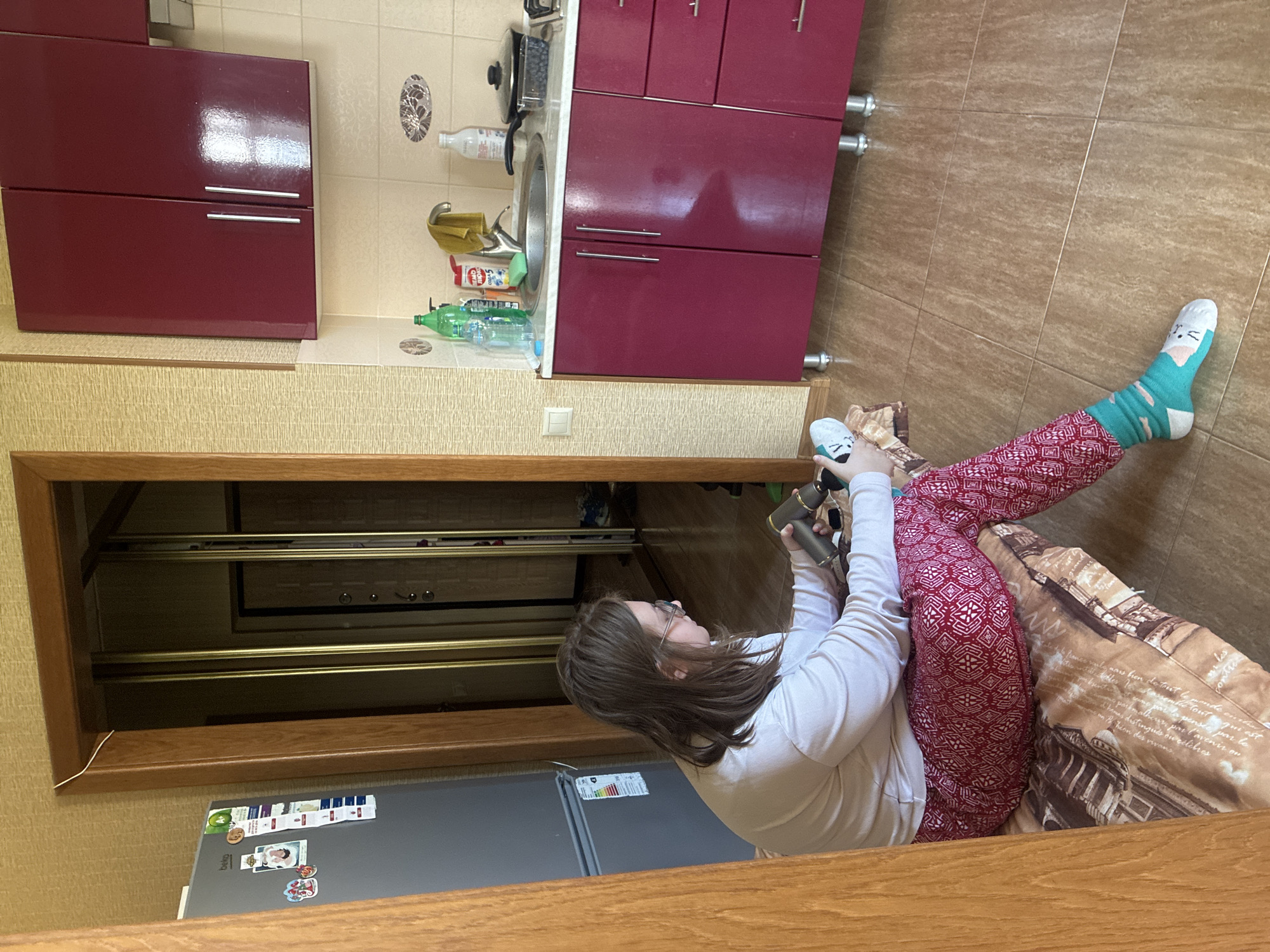 The image depicts an indoor scene, likely a kitchen or living area, within an apartment. A young woman with brown hair and glasses is seated on the floor, facing right. She is wearing a light-colored long-sleeved shirt, red patterned pants, and teal socks with cat designs. Her left hand is holding a cylindrical object, possibly a vacuum cleaner attachment, and she appears to be cleaning something on the floor. Her right hand is resting on her lap.
The environment includes a refrigerator on the left, a doorway with a mirrored closet beyond, and a kitchen counter with cabinets on the right. The kitchen counter features a sink, a faucet, various bottles and cleaning supplies, a pot with a lid, and a container of what appears to be blueberries. The flooring is tiled, and the walls are covered in a textured wallpaper. The overall impression is of a domestic setting. There is no visible indication that this location is in Sochi, Russia; it is a generic interior scene.