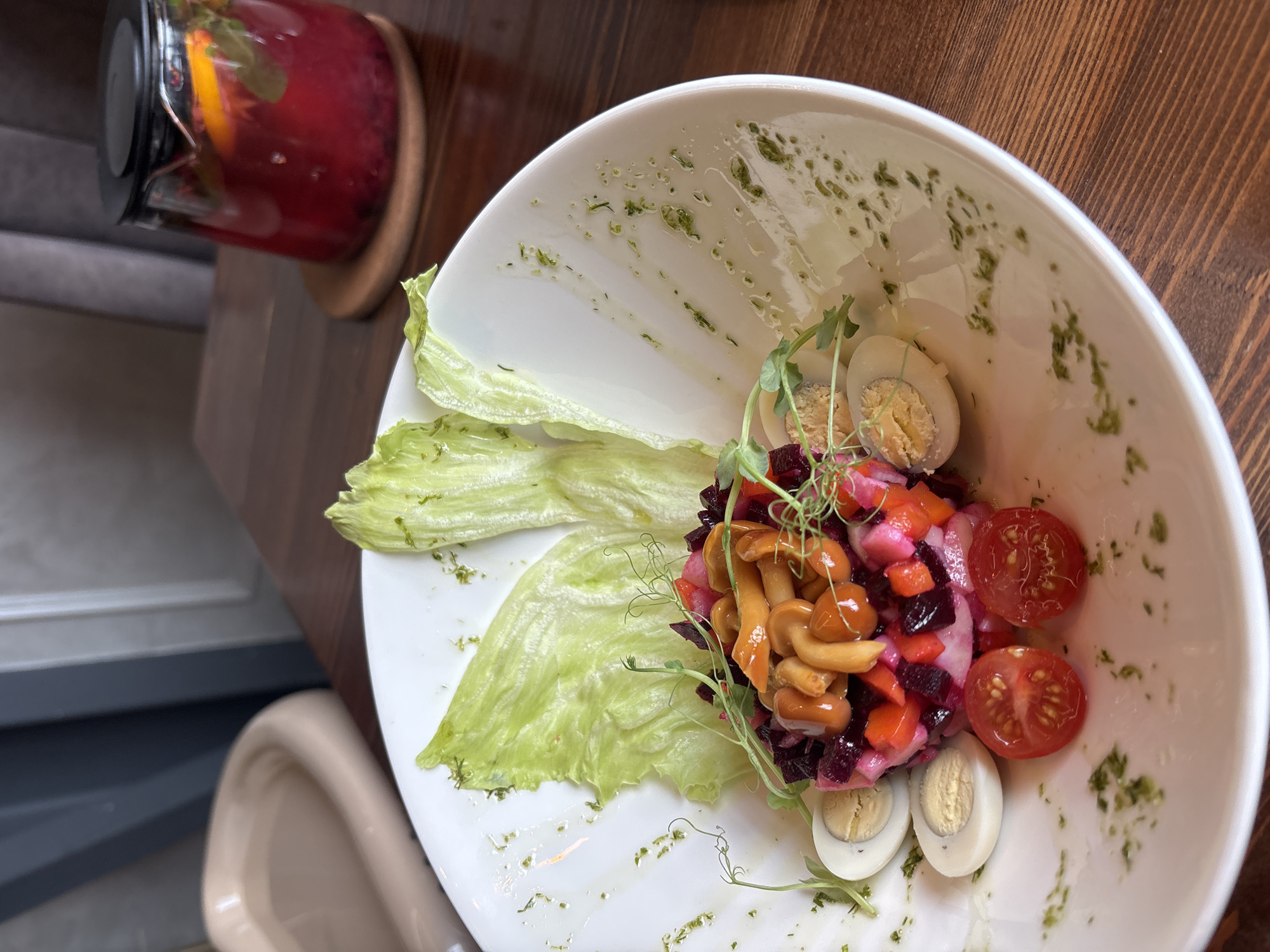 A close-up, high-angle shot shows a white plate of salad on a wooden table in a restaurant. The salad appears to be a beet-based salad with diced vegetables, mushrooms, halved quail eggs, and cherry tomatoes. Two large lettuce leaves are placed on the side of the salad. A glass teapot filled with a red beverage and garnished with orange slices and mint is visible in the background. The restaurant setting suggests it is located in Naberezhnye Chelny, Russia.