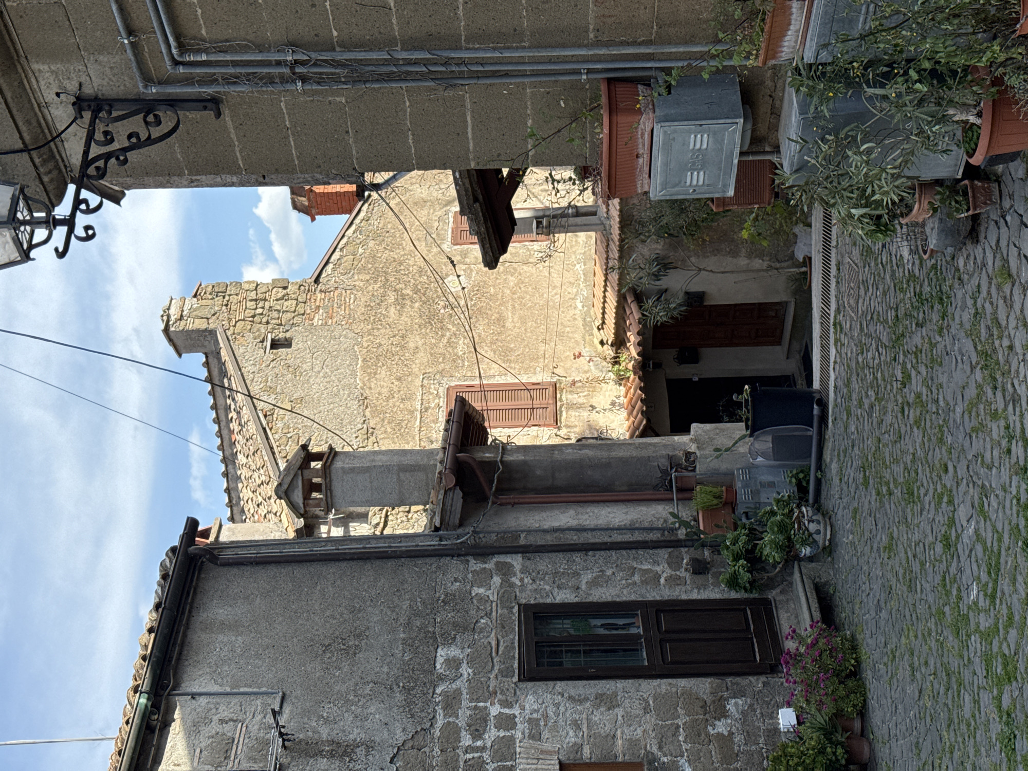 The scene is a narrow, angled view within the city of Castelnuovo di Porto, Italy. The foreground features a stone building with a dark-framed window, and potted plants with purple flowers are positioned at the base. Cobblestone pavement covers the ground, with grass growing between the stones. In the middle ground, another building displays weathered stucco, brickwork, and windows with shutters. A chimney and other architectural details extend above the roofline. The background reveals a glimpse of the sky with scattered clouds. Overhead, pipes and metal fixtures are attached to the buildings. The scene appears quiet and without human activity.