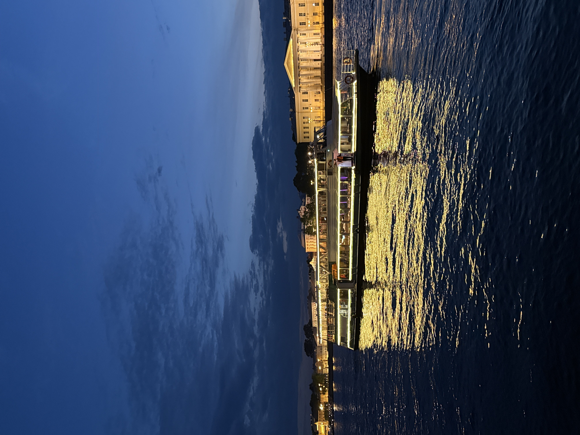 This image depicts a nighttime scene along a waterfront in Saint Petersburg, Russia, likely during twilight or early evening. The sky above is a deep, dark blue, with subtle cloud formations suggesting the fading light of day.
Along the embankment, a series of classical architectural buildings are visible, illuminated by warm, yellow lights. One prominent structure features a large triangular pediment supported by columns, characteristic of neoclassical design. Its windows glow, adding to the urban nighttime ambiance.
On the dark water, which appears to be a river or canal, a passenger boat is brightly lit. The boat's various decks and interior spaces are outlined with numerous yellow lights, creating a festive and inviting atmosphere. Several people can be discerned on board, presumably passengers enjoying an evening cruise. The bright lights from the boat, and potentially from the shore, cast long, shimmering golden reflections across the rippling water, creating a stark and beautiful contrast against the dark surface. The water itself shows texture from gentle movement, enhancing the play of light.
The scene conveys a sense of peaceful urban beauty, showcasing illuminated architecture and a lively boat tour against the tranquil backdrop of the night sky and water. No discernible text is visible within the image.