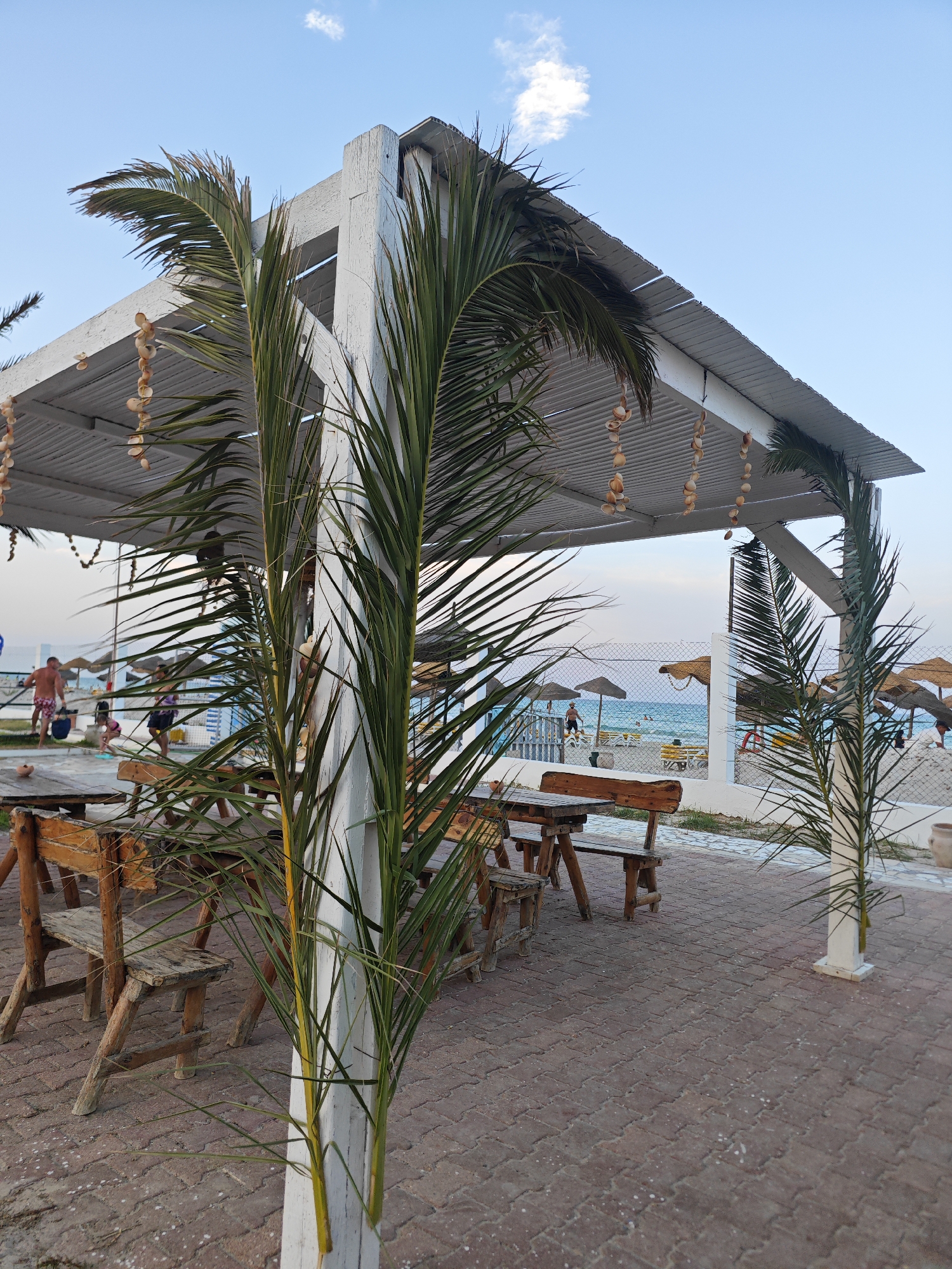 The image depicts a beachside setting in Mahdia, Tunisia, featuring a white wooden pergola adorned with strings of seashells. In the foreground, two palm fronds partially obscure rustic wooden picnic tables and benches set on a paved walkway. Beyond the pergola, the turquoise waters of the Mediterranean Sea are visible, with a sandy beach populated by a few individuals and umbrellas. The sky is a pale blue with a few wispy clouds. The scene suggests a relaxed atmosphere typical of a beach resort area.