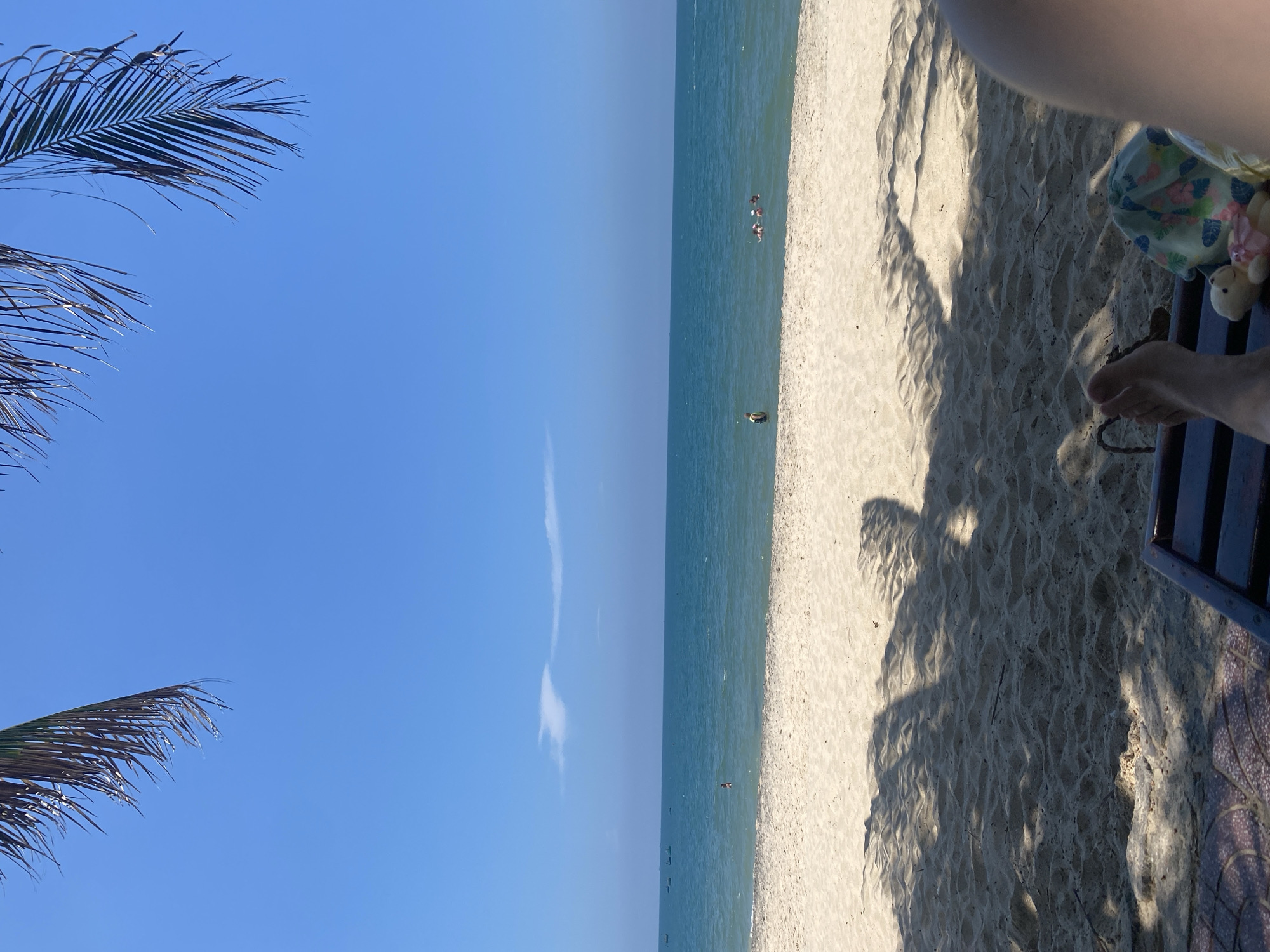 The image depicts a serene beach scene in Phu Thuy Ward, Vietnam. The foreground is dominated by a sandy beach with the shadow of palm fronds cast across it, suggesting a sunny day. A beach chair is partially visible in the lower right corner, with a person's legs resting on it, along with a small teddy bear. In the middle ground, the turquoise ocean stretches out to the horizon under a clear blue sky with a few wispy clouds. Several small boats are visible in the distance near the shore, and a few people are in the water, swimming or wading. The overall atmosphere is one of relaxation and a peaceful vacation.