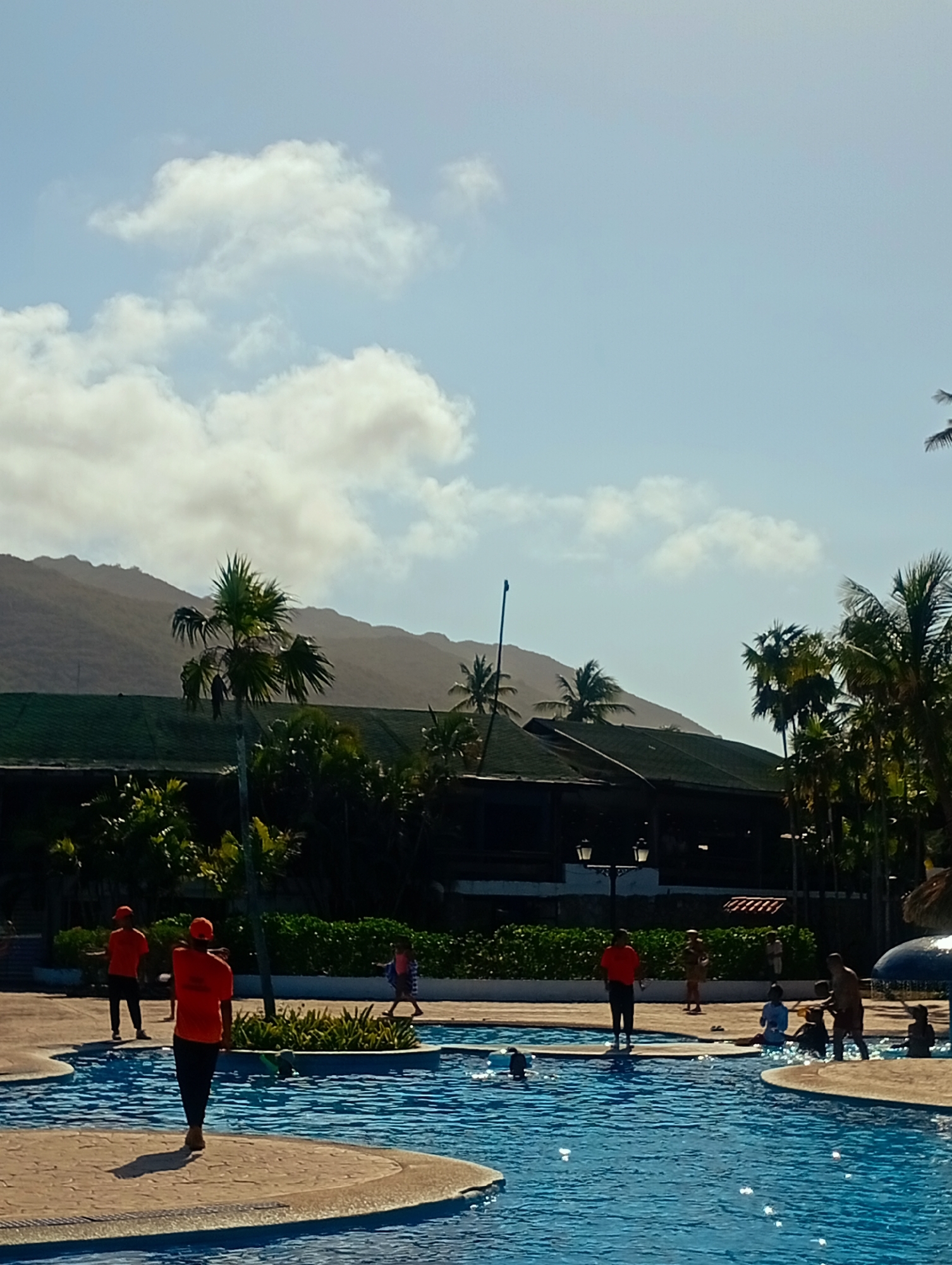 Playa El Agua, Venezuela. An outdoor swimming pool area is populated with several individuals engaged in various activities. Two people wearing orange shirts and dark pants stand on the pool deck, one walking and the other standing still. A child is also walking on the deck. Several other people are in the pool, some standing, others seated in the water. The pool has a paved surrounding area. Behind the pool is a building with a dark green roof and tropical foliage. In the background are hills, a partly cloudy sky, and some palm trees.