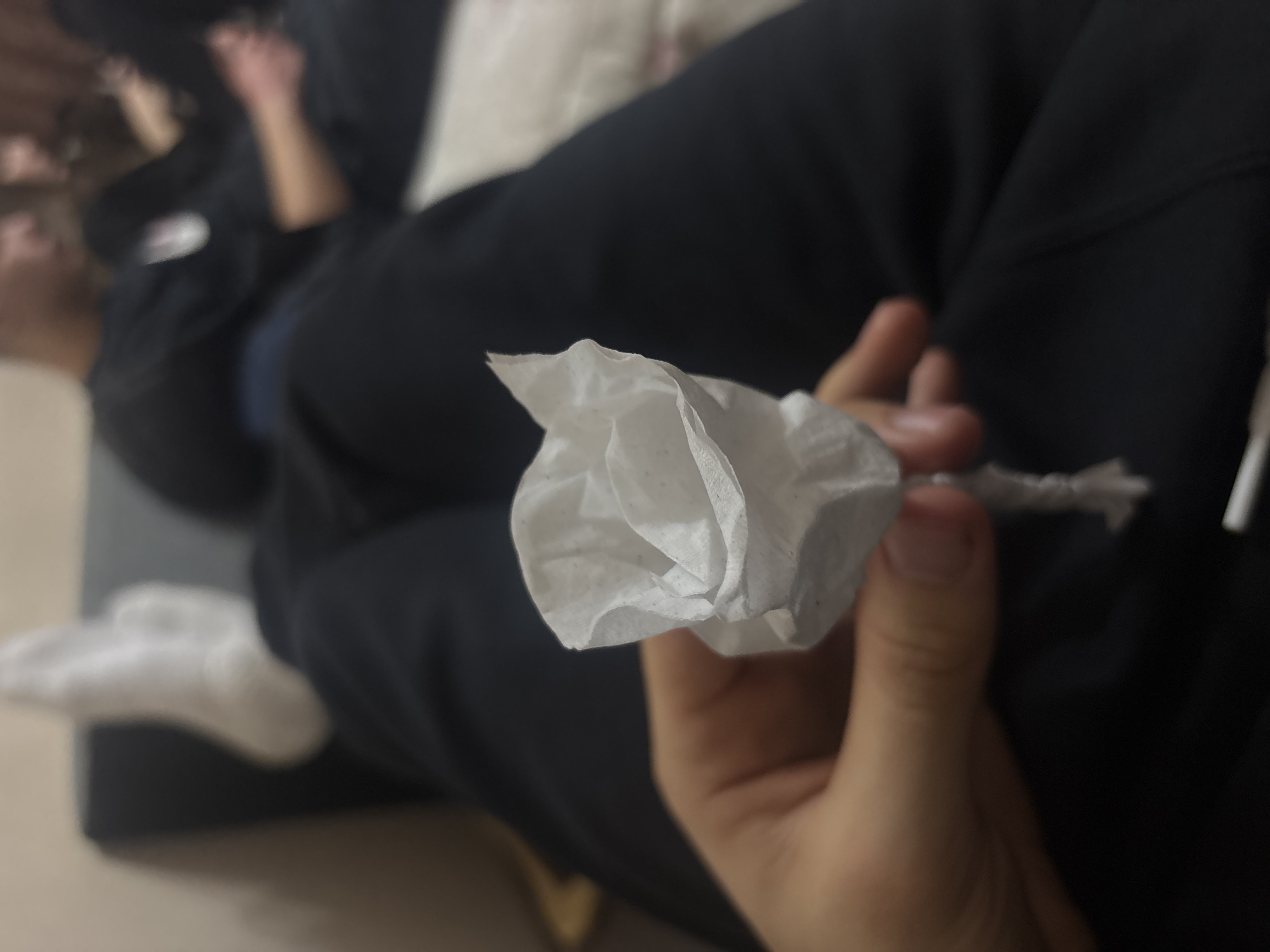 A close-up, angled shot shows a person holding a crumpled tissue in their hand. The tissue is twisted to resemble a flower. The person is wearing black sweatpants and white socks, and is sitting on a dark grey sofa. Another person, wearing a black zip-up sweatshirt, is partially visible in the background. The location is not possible to determine from the image.