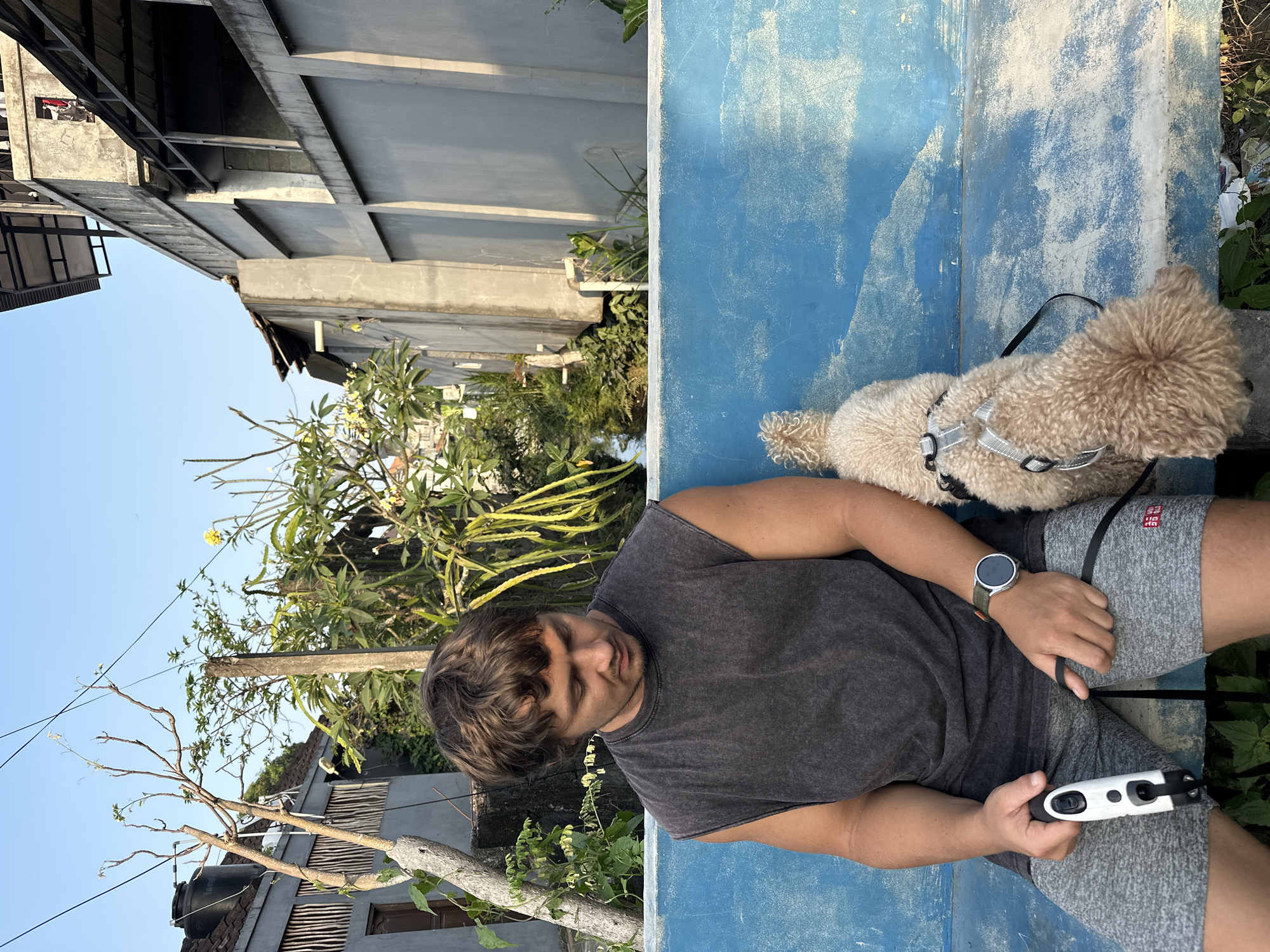 In North Kuta, Indonesia, a young man with brown hair, wearing a dark gray sleeveless t-shirt and gray shorts, sits on a weathered blue bench. He is looking down, holding a white dog leash in his left hand. A small, light-brown poodle mix dog, wearing a silver harness and black leash, sits next to him, also looking down. The background reveals a narrow waterway or canal flanked by dense green vegetation and trees. Beyond the greenery, the scene shows a mixture of residential structures, including concrete buildings with visible electrical wires, and some appearing under construction. The lighting suggests it is either early morning or late afternoon, with the sun casting long shadows.