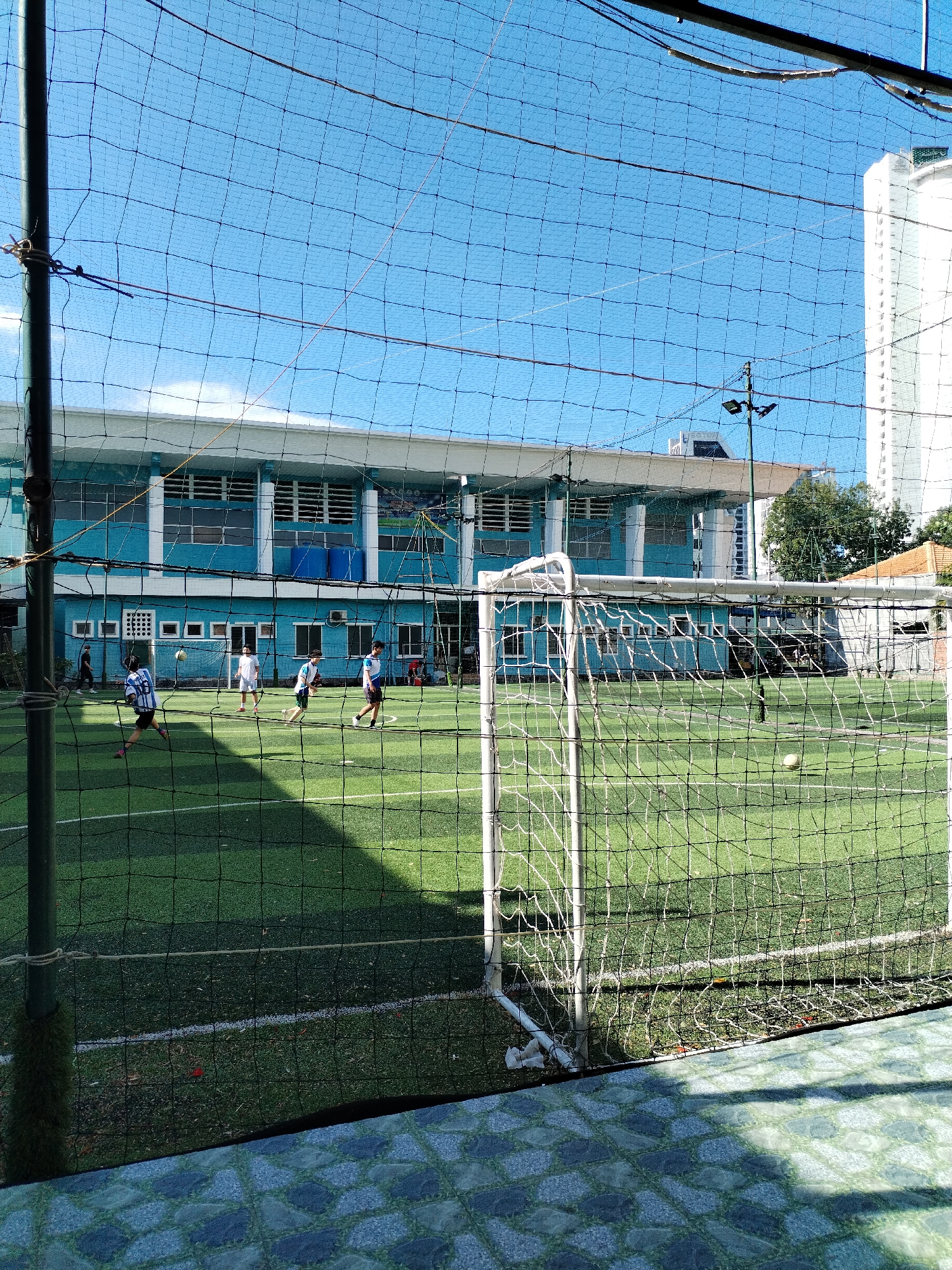 The image depicts a football match in progress on an artificial turf field. The field is enclosed by netting and a white-painted goalpost is in the foreground. Several players are engaged in the game, wearing light-colored jerseys and shorts. In the background, there is a light blue, two-story building with windows and a white roof. Behind the building, a tall, white high-rise building is visible against a blue sky. The photo was taken in Bac Nha Trang Ward, Vietnam.