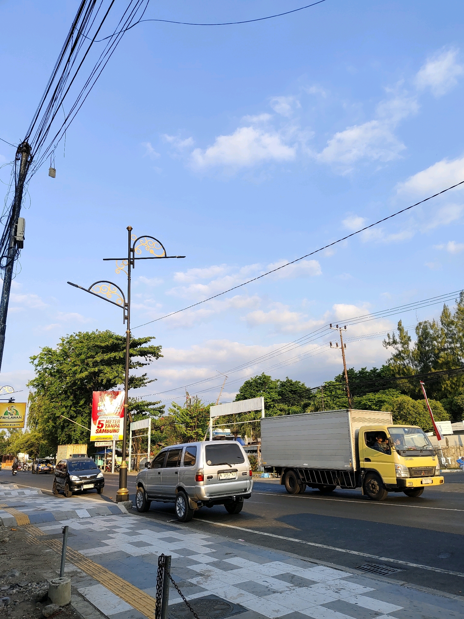 The image depicts a street scene in Probolinggo, Indonesia. In the foreground, a pedestrian walkway with a distinctive grey and white checkered pattern leads to the street. A silver SUV is parked on the side of the road, facing away from the camera. Further down the street, a yellow box truck with a driver visible inside is moving, accompanied by a black car in the adjacent lane. Overhead, a tangle of power lines stretches across the frame against a clear blue sky with scattered clouds. Decorative street lamps with curved, ornate tops stand along the sidewalk. In the background, lush green trees and several buildings line the street, indicating an urban or semi-urban environment. A red billboard with white text is also visible, advertising a printing service. The overall impression is a typical street view in Indonesia during daylight.
