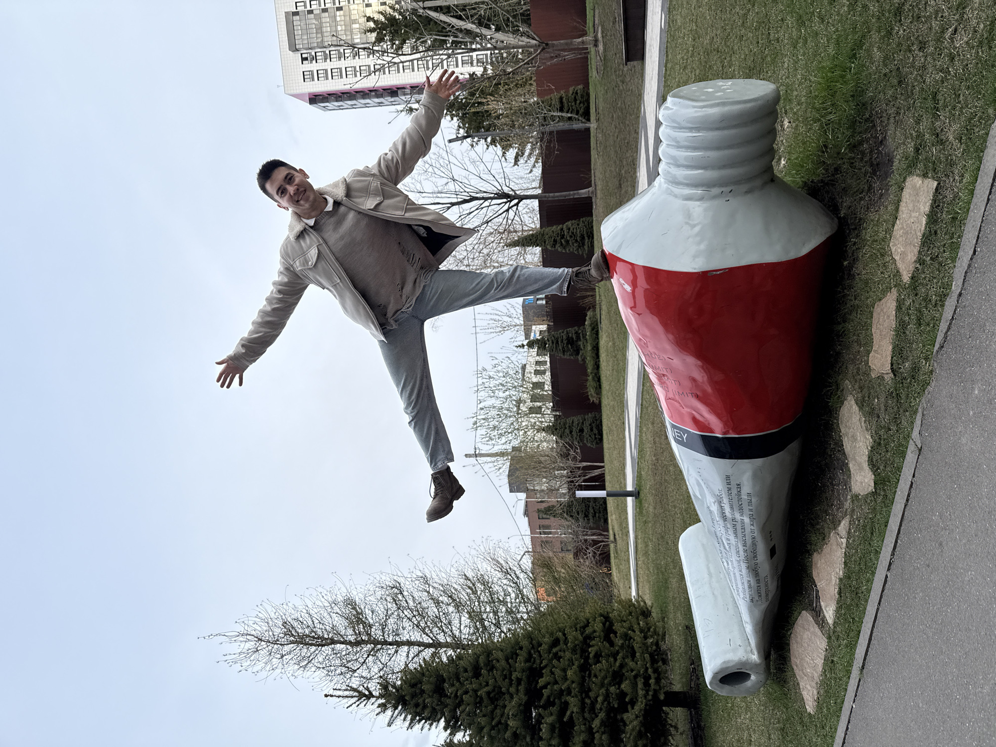 This media file, originally oriented vertically, captures a lively outdoor scene in an urban park in Kazan, Russia. A young man is depicted mid-jump, arms and legs extended outward, sporting a joyful smile. He wears a light-colored jacket over a grey sweater, grey jeans, and brown boots. His right foot is positioned near a large sculpture, indicating a playful interaction.
The prominent object is a colossal, horizontally placed art sculpture resembling a tube of paint. It is white with a distinct red section, capped by a grey screw top, and rests on green grass. Inscribed on its surface is Russian text detailing paint properties: "допративно-прикладных и графических работ. Разбавляется водой, специальным разбавителем или акриловым лаком. После высыхания водостойкая. Ложится на любую свободную от жира и пыли поверхность." Additionally, fragments like "MIT," "NO IM," "EY," and the number "19470" are visible.
The background features an overcast sky, suggesting daytime without direct sunlight. Various trees, including both bare deciduous and dark green evergreens, are scattered around, contributing to the park-like atmosphere. Modern multi-story buildings stand in the distance, indicating an urban setting. A paved path runs alongside the grassy area. The man's dynamic pose and cheerful expression convey a sense of fun and spontaneity.