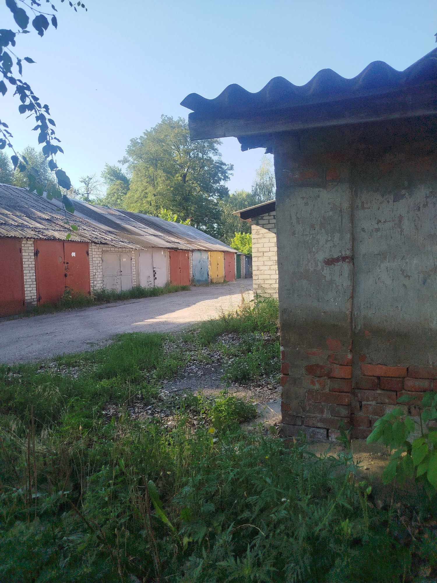 The image depicts a row of brick garages with corrugated roofs under a clear blue sky. The garages are situated on a dirt road, and the surrounding area is overgrown with grass and trees. The garages appear to be old and somewhat dilapidated. The front of the garages are painted in various colors, including red, white, yellow, and blue. Some of the garage doors have numbers or markings on them. In the foreground, there is a wall made of brick and concrete. The scene appears to be a typical garage complex in a residential area. The specific location is a garage complex in Kremenchuk, Ukraine.