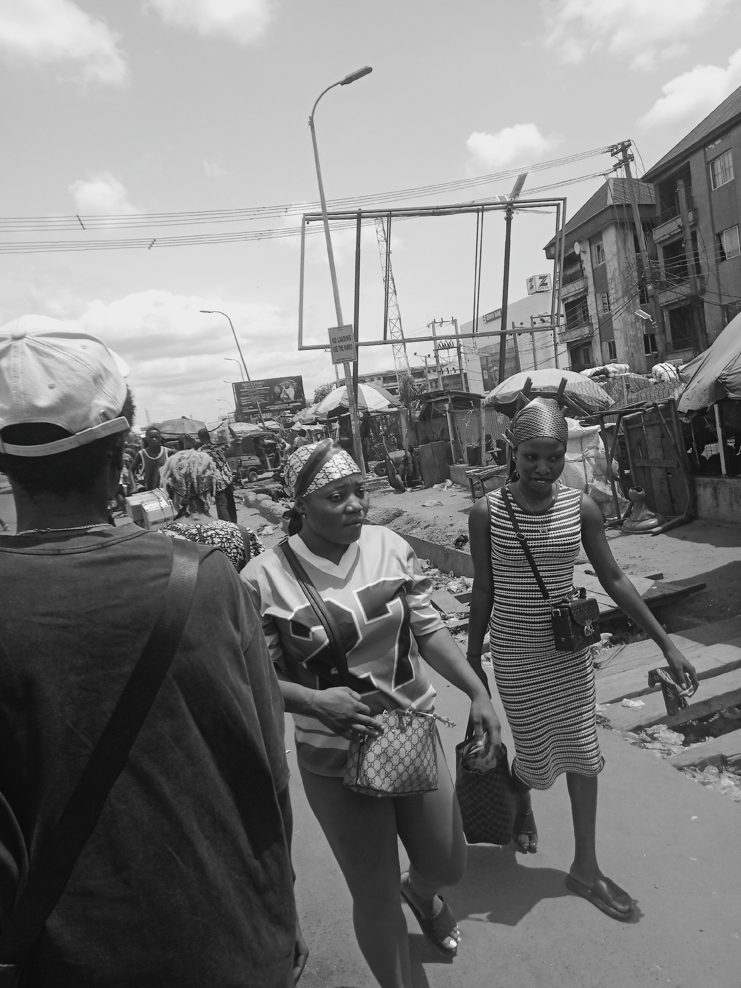 The black and white photograph depicts a bustling street scene, likely a market or commercial area in an urban environment. The image is captured from a pedestrian's perspective, with several individuals moving through the foreground and middle ground.
In the immediate foreground, the back of a person wearing a light-colored baseball cap and a dark t-shirt is visible. To their right and slightly ahead, a woman wearing a patterned headscarf and a sports jersey (with a partial number "22" or "25" visible) walks with a patterned handbag, looking towards the right side of the frame. Further to her right, another woman walks forward, also adorned with a patterned headscarf, a horizontally striped sleeveless dress, and carrying two bags. She appears to be smiling faintly. Other figures are present in the mid-ground, navigating between market stalls and structures.
The background features a complex network of overhead power lines, streetlights, and a large, empty billboard frame dominating the center. Buildings of varying heights line the street; one building clearly displays the "ZENITH BANK" logo. A white sign attached to a street pole on the right side of the frame reads, "NO LOADING USE THE PARKS." The ground appears somewhat uneven and unpaved in places, with visible debris consistent with a busy street or market. The sky is bright with scattered clouds, indicating a daytime setting, likely sunny, given the visible shadows. The overall scene suggests a vibrant, active street life.