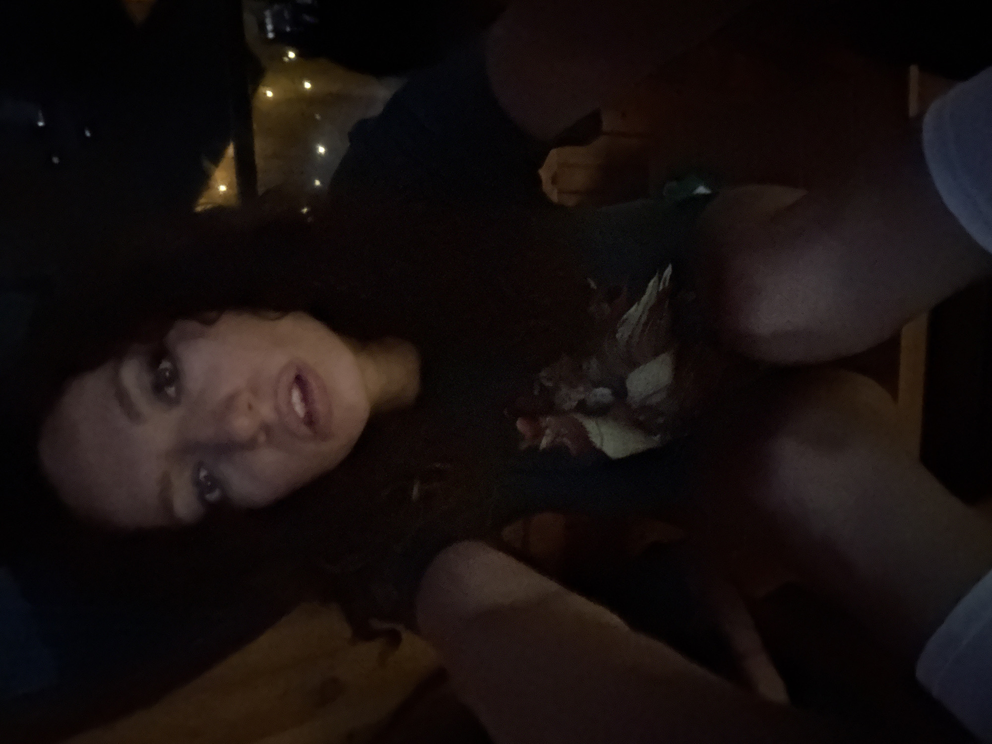 A woman with long, dark hair is lying down and looking at the camera. She is wearing a dark-colored shirt with a printed design. The environment appears dimly lit, possibly indoors, with string lights visible in the background. The image also contains parts of other people’s bodies: a light-skinned arm and leg or thigh. The location of Bắc Hà, Vietnam, cannot be confirmed from the visual content alone.