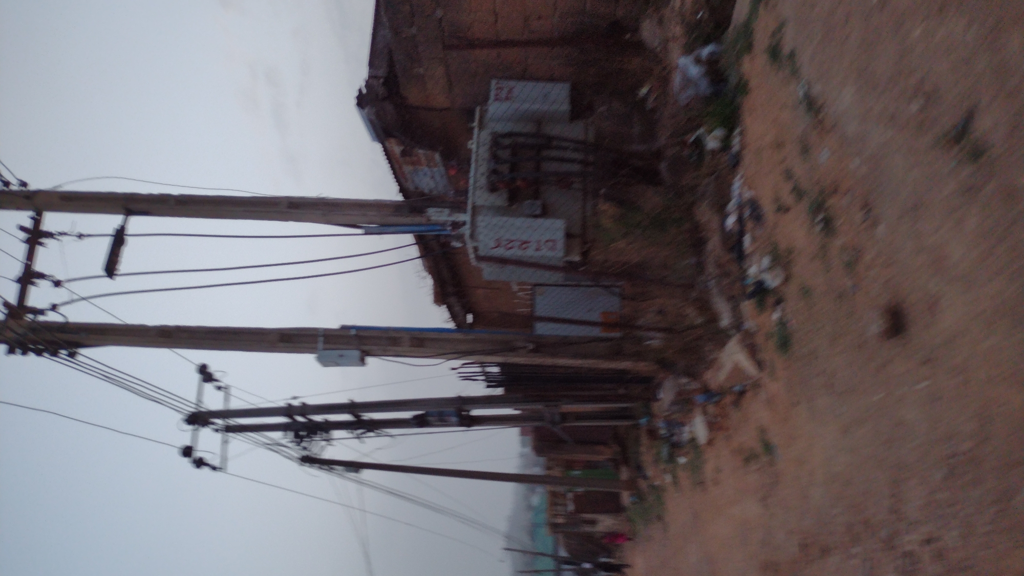 The image depicts a street scene in Jos, Nigeria, characterized by utility infrastructure and roadside conditions. Several wooden utility poles support an array of power lines. A transformer is mounted on a pole near a building with a corrugated metal roof. Trash and debris are scattered along the roadside. The environment is overcast, and the overall impression is one of a developing urban setting.