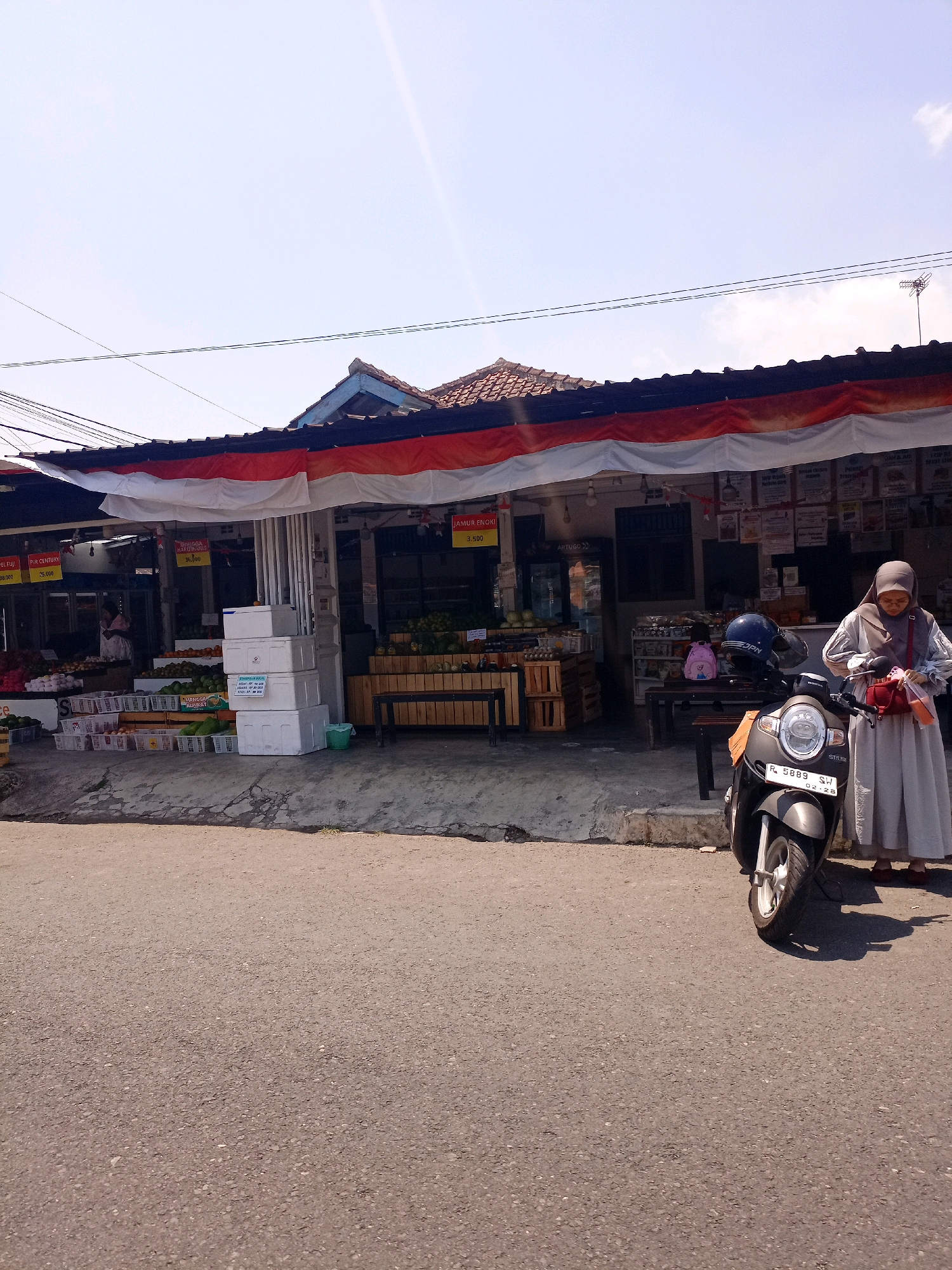 A street-level view captures a small market stall in Banjarnegara, Indonesia. The stall, adorned with Indonesian flag colors, displays a variety of produce in baskets and crates. A woman in a headscarf stands beside a parked motorcycle, rummaging through a bag. The scene is bathed in bright sunlight under a clear sky.