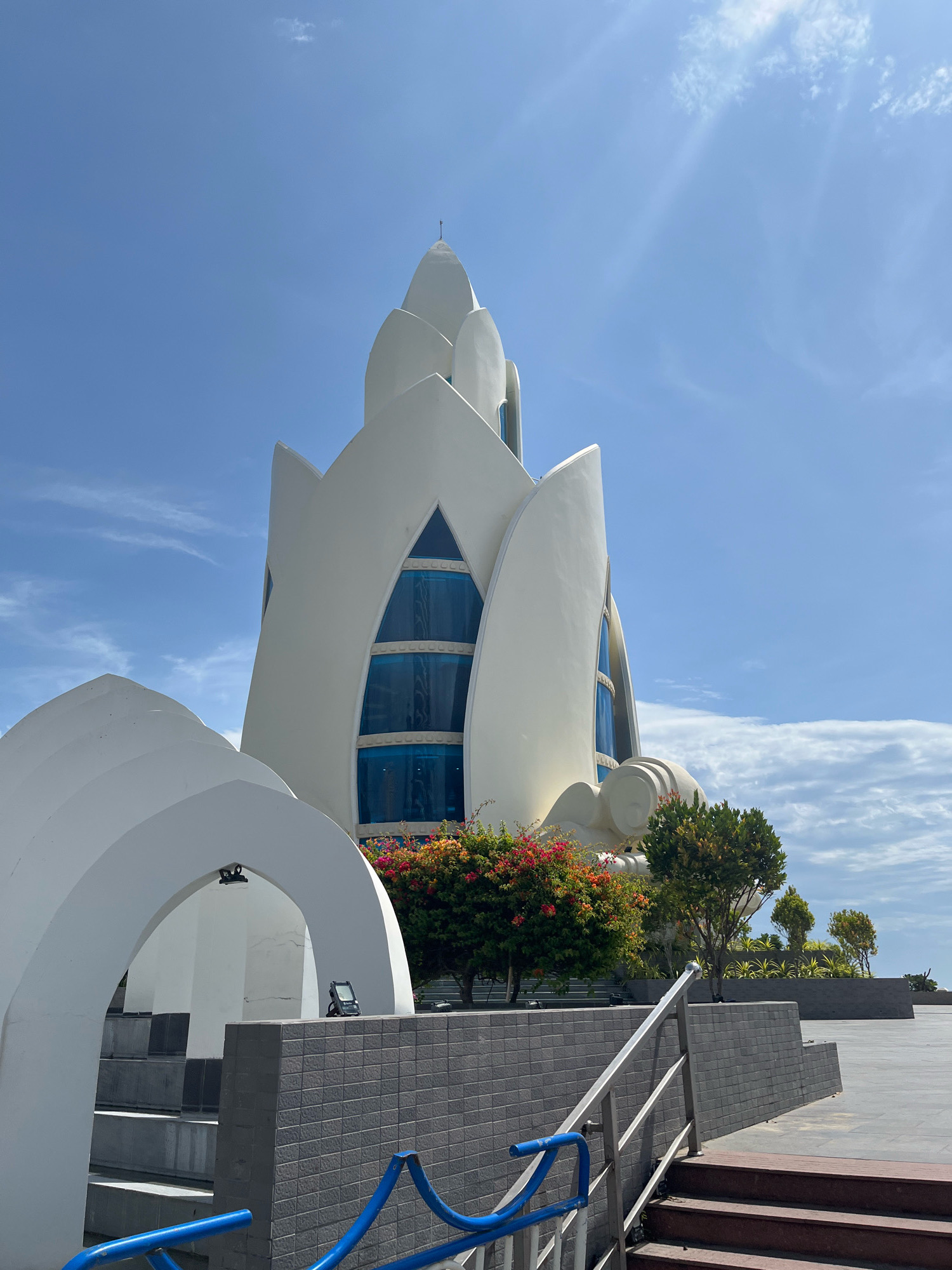 This is a photograph of the Long Son Pagoda in Nha Trang, Vietnam. The iconic white, lotus-shaped building dominates the frame, its smooth, curving white walls reaching towards a bright blue sky dotted with wispy clouds. Large, triangular blue-tinted windows are visible within the structure. In the foreground, a white arched entryway leads to a set of grey steps. To the right of the steps, a dark grey tiled wall with a blue metal handrail frames a section of the plaza. Lush green trees and vibrant red and orange flowers are visible around the base of the pagoda. The overall impression is one of a modern, architecturally striking religious site on a clear, sunny day.