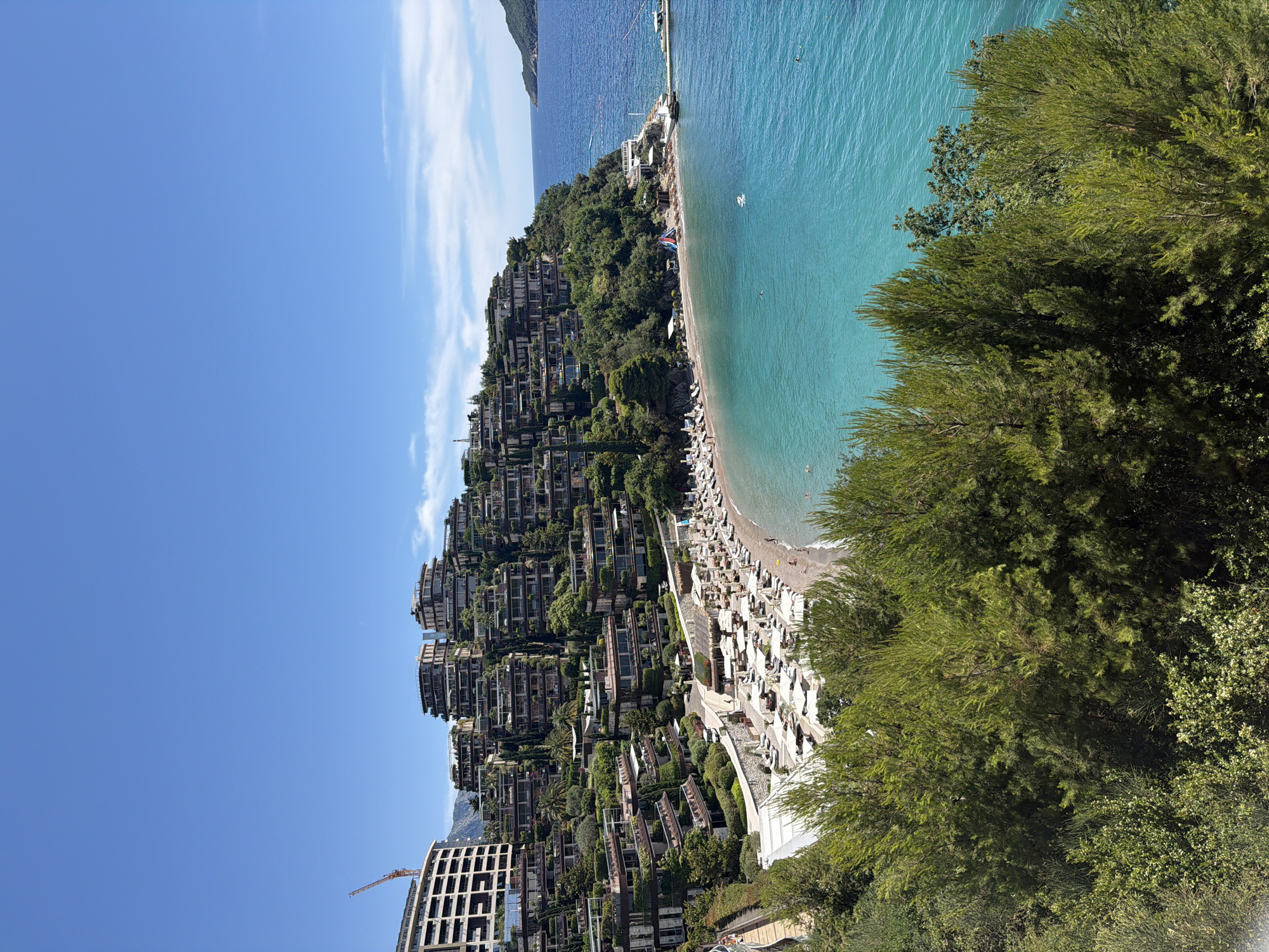 This is a high-angle, wide shot of a resort complex situated on a hillside overlooking a crescent-shaped beach and a turquoise sea. The resort consists of multiple terraced buildings with modern architecture, each featuring balconies and abundant greenery. The beach is lined with numerous white lounge chairs and umbrellas. The sea is calm, with a few people swimming and a small boat docked at a pier. The sky is a clear blue with a few wispy clouds. The resort appears to be located in the vicinity of Budva, Montenegro.