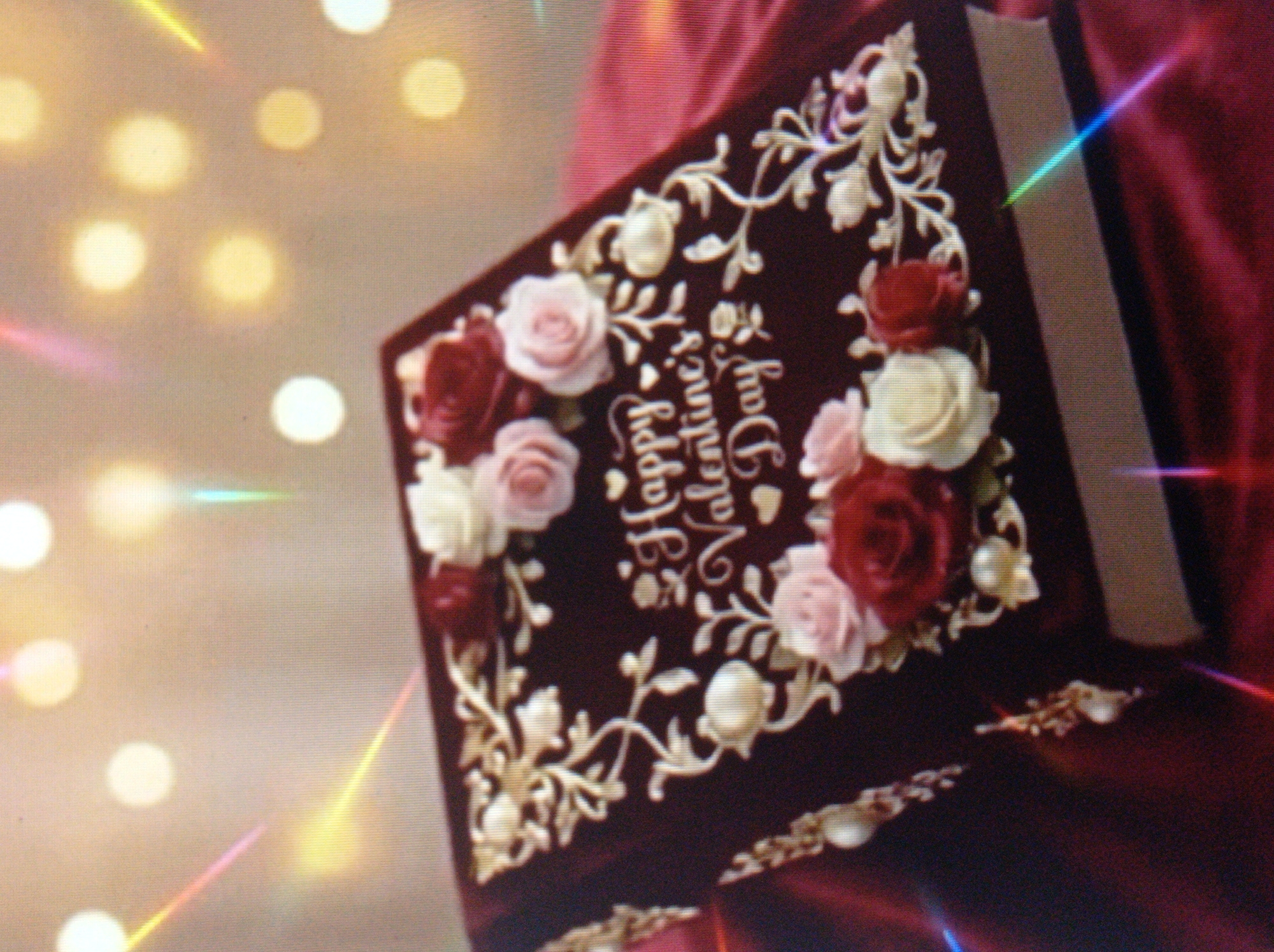 The image shows a close-up of a dark red, book-shaped box, possibly a gift or candy box, decorated with intricate gold floral scrollwork and artificial roses in shades of pink, white, and deep red. Some of the roses appear to be made of fondant or similar edible material, and there are small pearl-like embellishments scattered among the floral designs. The text "Happy Valentine's Day" is written in a cursive white font on the lid of the box, with small heart shapes interspersed. The box is placed on a dark red, satin-like fabric. In the background, there are soft, out-of-focus circular lights, creating a bokeh effect, suggesting an indoor setting with decorative lighting. The image is slightly blurry and has some lens flare effects with streaks of light in various colors. There are no people or animals visible in the image. The image does not contain any information that would allow it to be located in the city of Mukachevo, Ukraine.