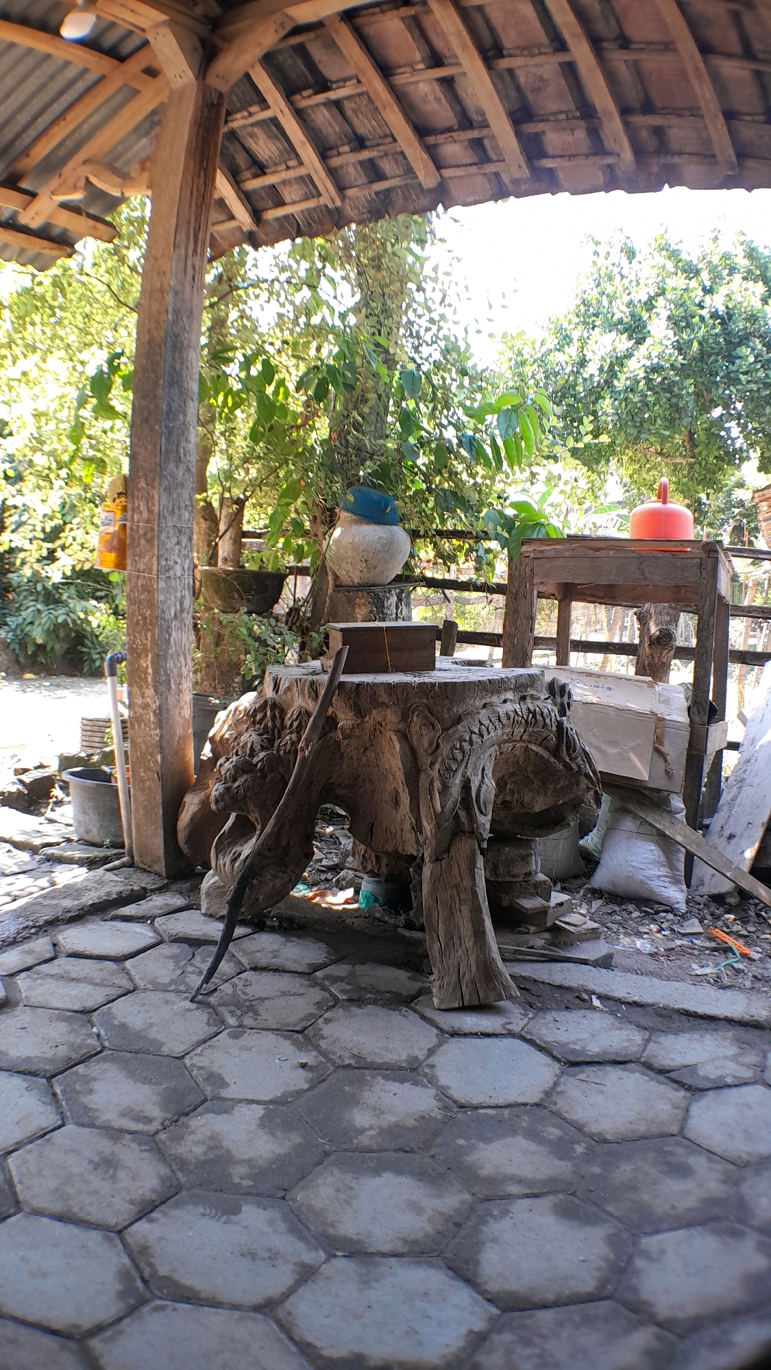 An outdoor scene is depicted in Sumberarum, Indonesia, under the partial cover of a weathered wooden structure with a corrugated metal roof. In the foreground, hexagonal paving stones are wet and stained. A large, intricately carved wooden stump, resembling a mythical creature with detailed facial features, serves as the central object. A thin, dark wooden pole leans against it, pointing towards the ground. On top of the stump rests a small wooden box with a string tied around it. To the right, a rustic wooden table supports an orange kettle and some cardboard boxes. Behind the table, a white bag and other debris are visible. Further back, a large, light-colored pot with a blue lid sits on a raised platform. The environment is lush with green foliage from trees and vines, some of which are growing against a wooden fence. Sunlight filters through the leaves, creating dappled light and shadows on the ground. The overall impression is one of a rustic, possibly outdoor workshop or storage area.