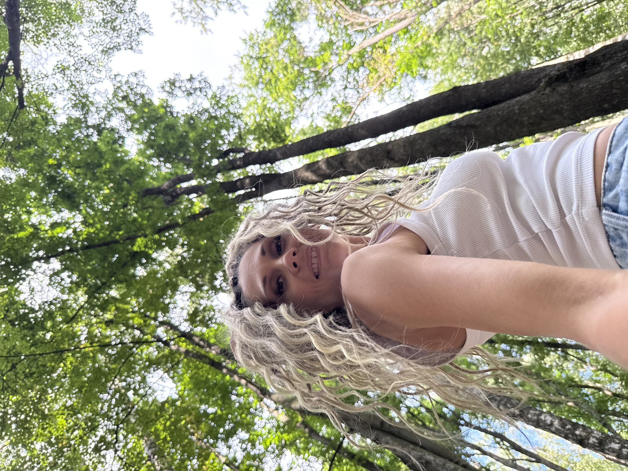 A low-angle, wide shot captures a woman with blonde, curly hair posing in a forest. She is wearing a white tank top and blue jeans. The woman is looking up and to the right, with a slight smile. She is positioned in the foreground, with tall trees and green foliage filling the background. The sunlight filters through the leaves, creating a dappled effect. The location is identified as Rusanovo, Russia.