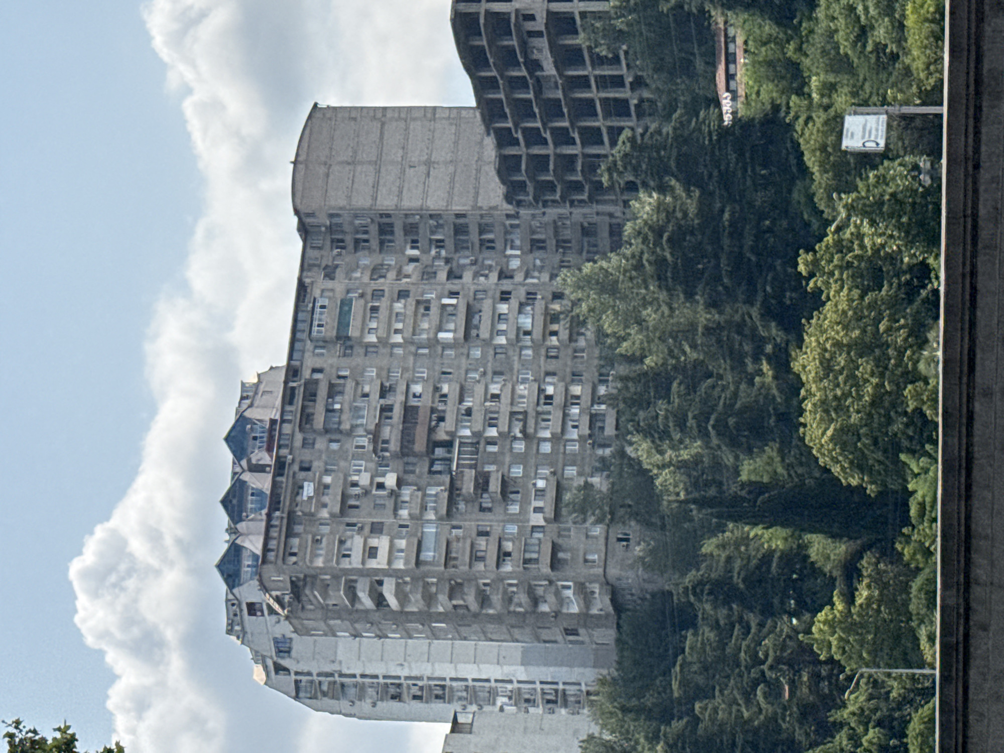 The image captures a view of several apartment buildings and an unfinished construction project in Tbilisi, Georgia. The primary focus is on a large, grey, multi-story apartment complex with numerous balconies, characteristic of Soviet-era architecture. To its right, a partially constructed building with exposed concrete floors and columns is visible. The structures are situated on a hillside covered with dense green trees. In the foreground, a concrete barrier runs horizontally across the bottom of the image. The sky above is blue with scattered white clouds. A street light and a signpost are also discernible amongst the trees. The overall impression is an urban landscape with a mix of completed residential buildings, ongoing construction, and lush natural surroundings.