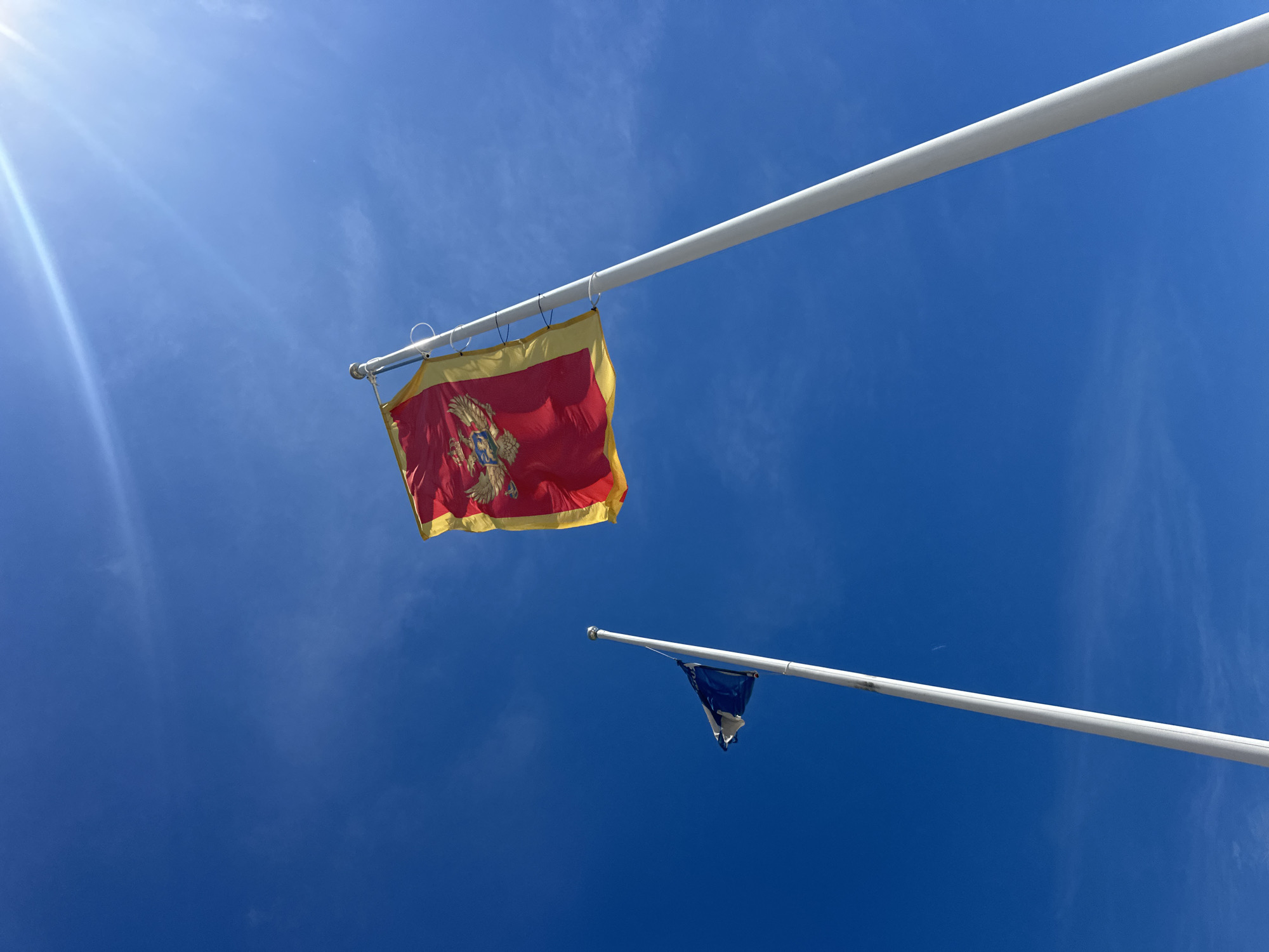 Two flags are flying on flagpoles against a bright blue sky with some wispy clouds and the sun glinting. The larger flag, in the foreground and to the right, is red with a gold border and features a prominent coat of arms in the center. This appears to be the flag of Montenegro. To the left and slightly behind it, a smaller flag is flying on a separate flagpole. This flag is dark blue with a circle of gold stars, characteristic of the European Union flag. The flagpoles are white and tall, with the one on the right extending higher than the one on the left. The scene is outdoors, under a clear sky. The presence of the Montenegrin and EU flags suggests a location of official significance, possibly a government building or international point of entry. The city of Boreti, Montenegro, is a coastal town on the Adriatic Sea, known for its beaches and tourist resorts, and this image could be taken at such a location.