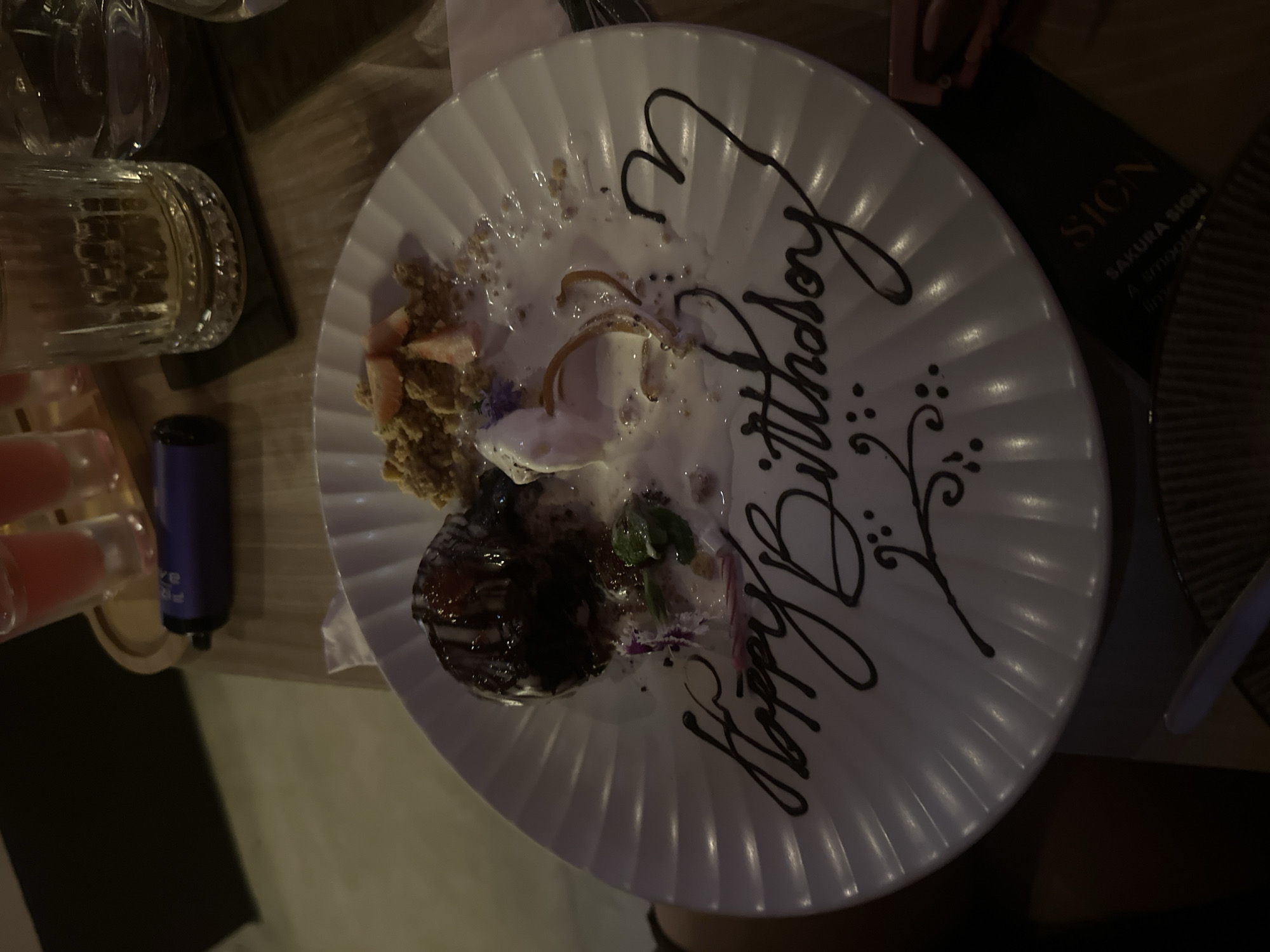 A partially eaten dessert cake sits on a white plate with "Happy Birthday" written in chocolate sauce. Visible remnants of the cake include crumbly textures, fruit pieces (possibly strawberries), a scoop of melted cream, a chocolate-covered element, and some greens. To the left of the plate, there's a vape pen and a clear glass with a beverage. The plate rests on a table; a menu with "Sakura Sign" is partially visible. The location is North Kuta, Indonesia. No people are fully visible in the frame.