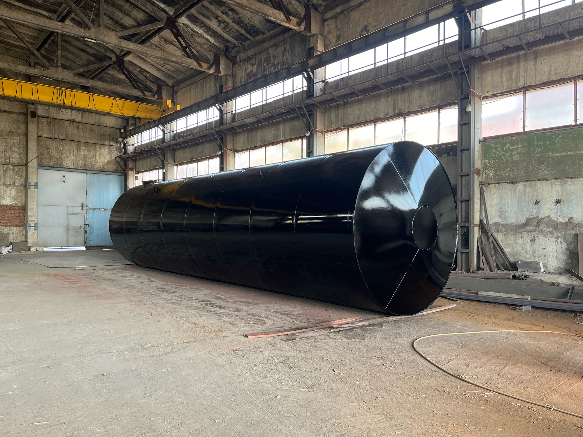 A large, black, cylindrical industrial tank lies horizontally on the concrete floor of a large, open, industrial building. The building has a high, corrugated metal roof supported by exposed beams and trusses. Natural light streams in through a series of windows high up on the walls. A yellow overhead crane is visible on the left side of the image. The tank is the dominant object in the scene, taking up a significant portion of the frame. The floor is concrete with some reddish dust or markings. In the background, on the left, are large industrial doors. On the right, there are piles of metal stock and building materials. The scene appears to be a workshop or manufacturing facility within the Taganrog City District, Russia, given the context. There are no people visible in the image.