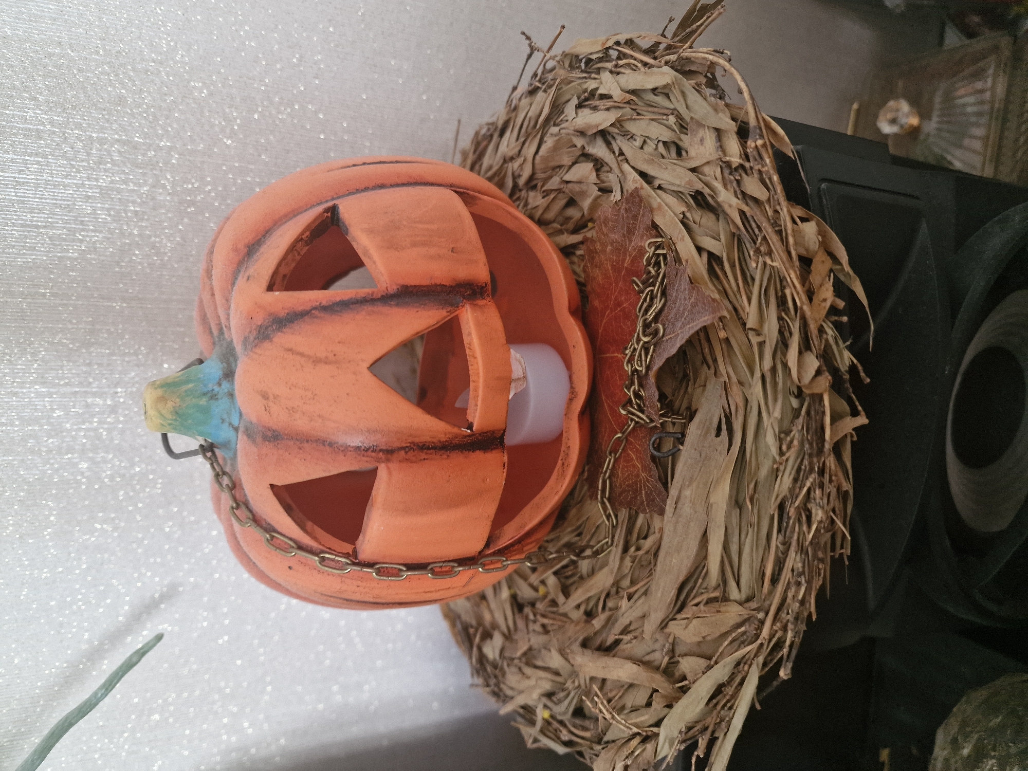 A ceramic jack-o'-lantern with a metal chain draped around it sits on a nest of dried leaves and twigs. A small white LED candle glows within the pumpkin. The background is a white, textured surface, possibly a wall. The scene is likely indoors, on a table or shelf. No specific location in Nuremberg, Germany is indicated by the image.