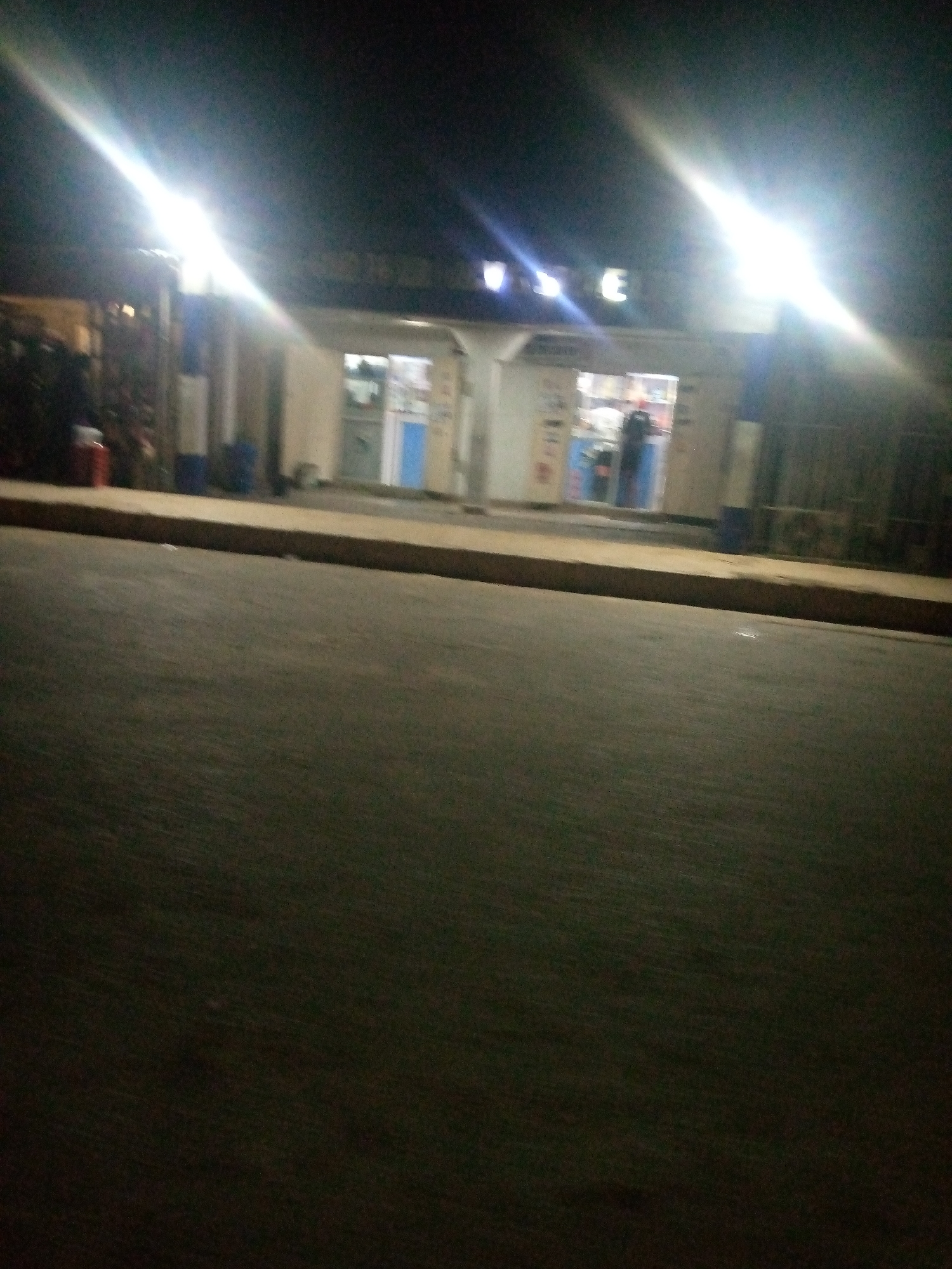 The image shows a blurry, nighttime scene of a storefront in Jalingo, Nigeria. Bright lights illuminate the building, which appears to be a shop with display windows. A person is visible inside the shop, standing behind a counter. The area in front of the store is paved, suggesting it is a public street or sidewalk. The overall impression is of a well-lit commercial establishment operating after dark.