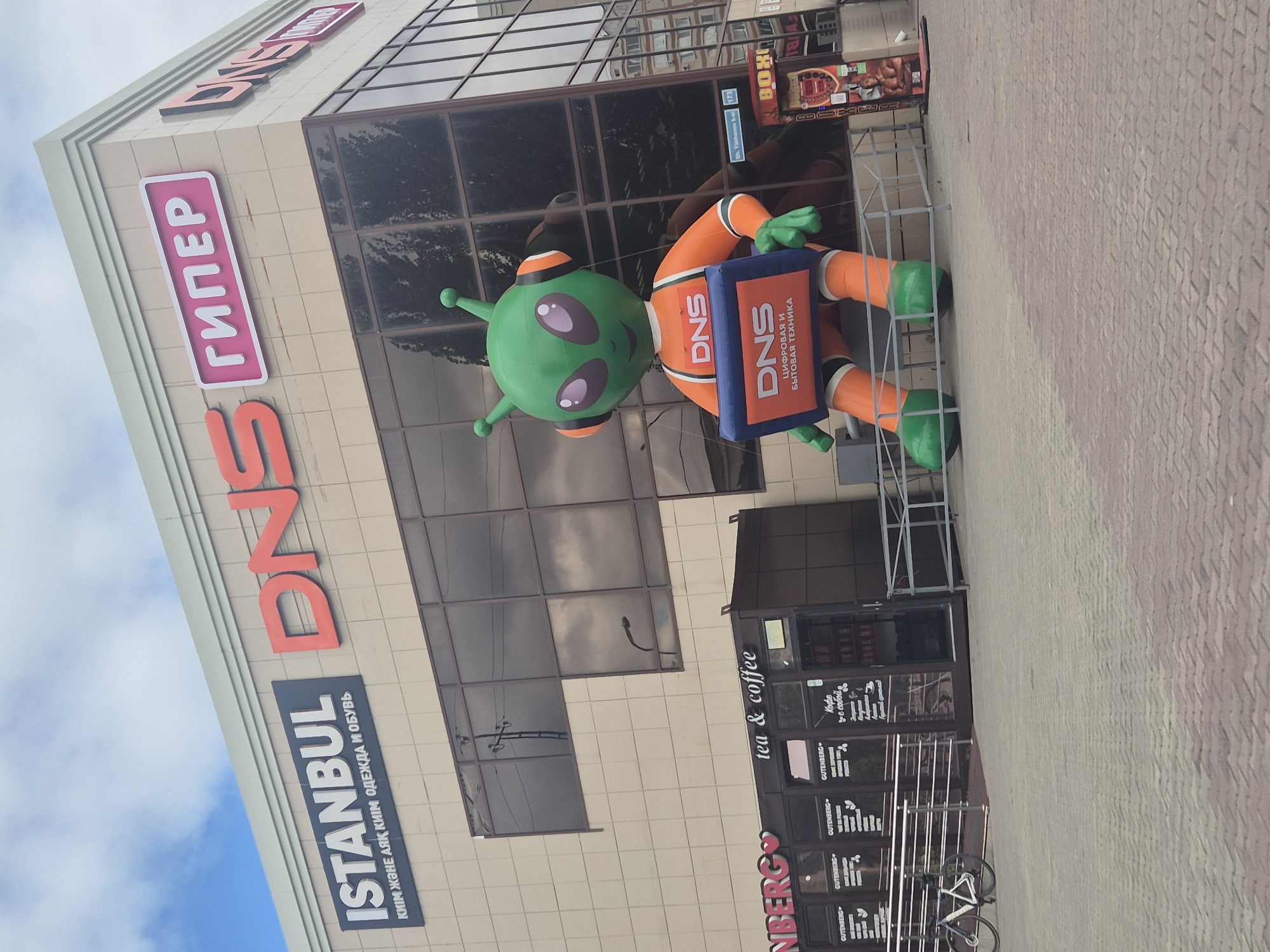 The image shows a large inflatable alien mascot in front of a building with multiple signs. The mascot is green with orange clothing and is holding a blue sign with "DNS" and "Цифровая и бытовая техника" (Digital and household appliances) written on it. Above the mascot, the building has signs that read "ISTANBUL" and "DNS ГИПЕР". Below the "ISTANBUL" sign, there's text in Kazakh that translates to "clothing and footwear". Another sign reads "tea & coffee". In the background, there is a bicycle parked near a railing. The building appears to be a retail establishment. The scene is an outdoor urban environment with a paved walkway. The sky is overcast. This location is identified as the DNS store at Sh. Ualikhanov St, 173, Kokshetau, Kazakhstan.