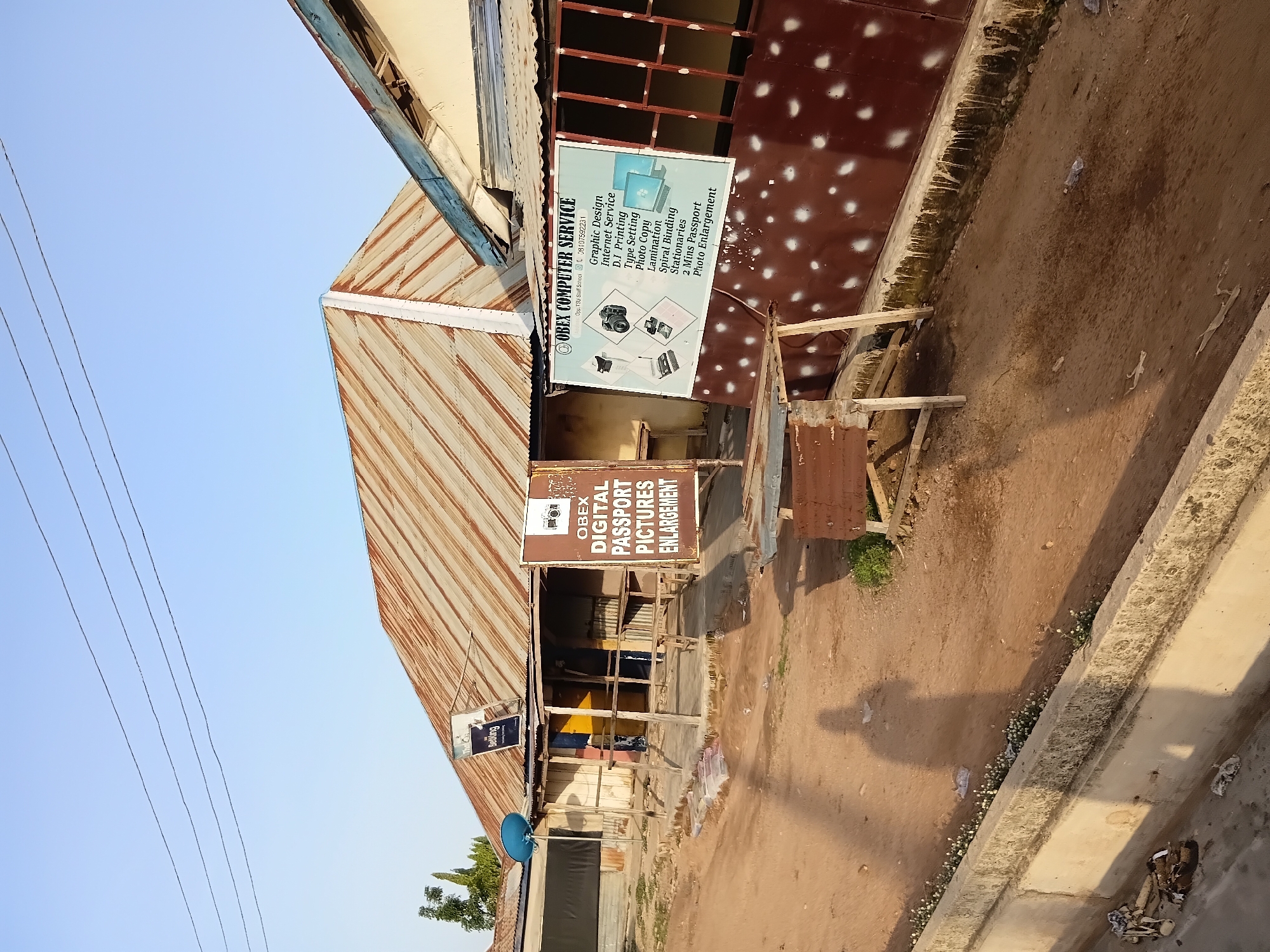 The image depicts a street scene in Jalingo, Nigeria. On the right, a commercial establishment with a corrugated metal roof and a dark brown facade sprayed with white dots advertises "OBEX COMPUTER SERVICE," listing services such as graphic design, internet service, D.I. printing, type setting, photo copy, lamination, spiral binding, stationaries, 2 mins passport photos, and photo enlargement. A sign above the entrance to the building on the left reads "OBEX DIGITAL PASSPORT PICTURES ENLARGEMENT." The structures appear weathered, with exposed wooden supports and rusty metal roofing. The ground is dirt and uneven. In the foreground, a concrete drainage channel runs along the bottom of the image, containing some scattered debris and small white flowers. Power lines are visible against a clear blue sky. The shadow of a person is cast across the dirt ground. A satellite dish is mounted on the roof of one of the buildings.