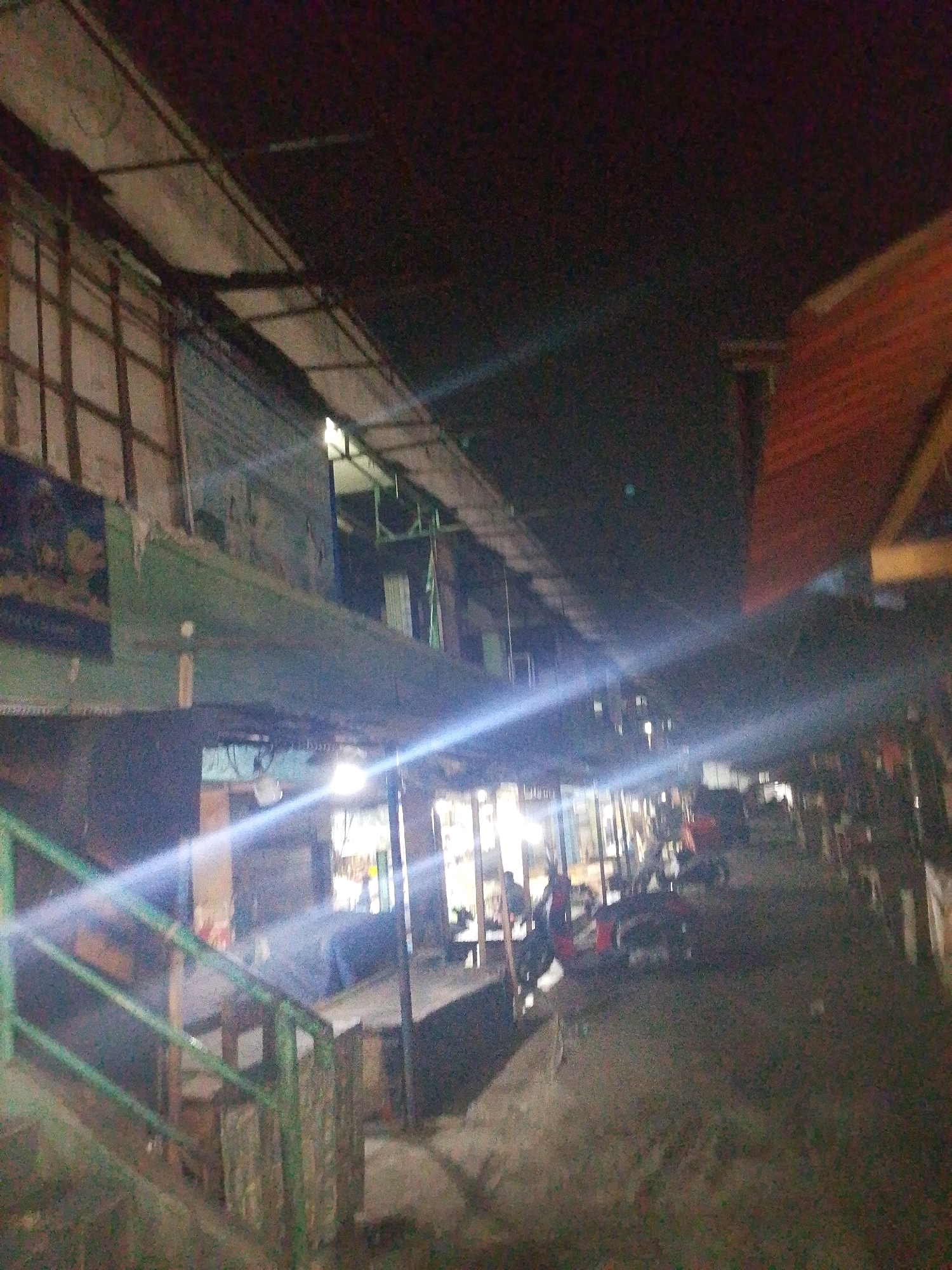 The image depicts an outdoor scene at night, likely a marketplace or a row of shops in Karawang, Indonesia. The lighting is dim, with strong artificial light sources creating prominent light streaks across the frame. In the foreground, a green railing is visible, suggesting an elevated walkway or a staircase. Beyond the railing, a covered walkway or stalls are lined up, with structures suggesting storefronts or seating areas. Several motorcycles are parked or are present in the scene, indicating a mode of transportation common in the area. The overall impression is one of a bustling, yet dimly lit, urban environment, characteristic of nighttime commerce in many Indonesian cities. The image quality is poor, with low resolution and noise, making it difficult to discern fine details.
