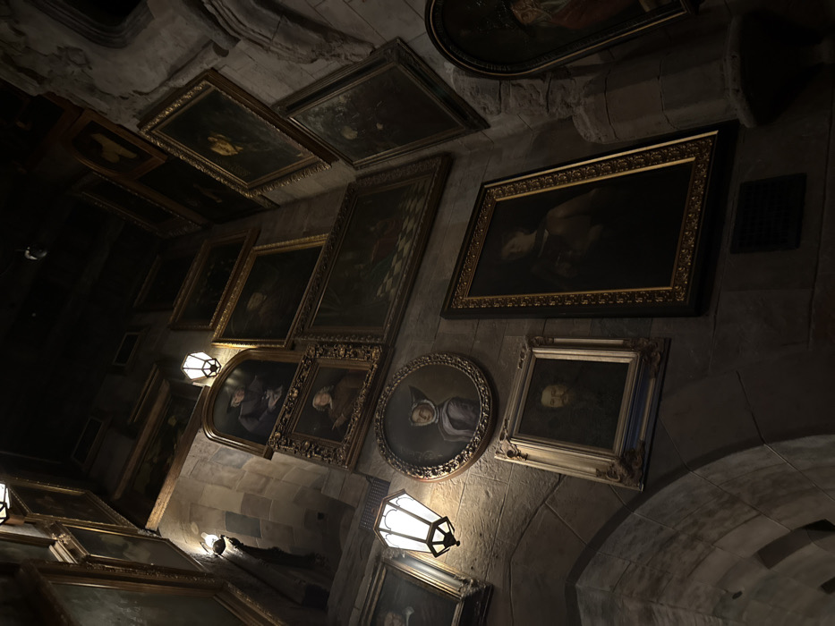 The image depicts a dimly lit interior chamber within The Wizarding World of Harry Potter - Hogsmeade, located at Universal's Islands of Adventure in Orlando, Florida, United States of America. This specific area is part of the queue for the "Harry Potter and the Forbidden Journey" attraction.
The scene features rugged, dark gray stone walls constructed from large, irregular blocks, suggestive of a gothic castle interior. The upper portions of the walls and ceiling are shrouded in deep shadow. Illumination is provided by several wall-mounted, lantern-style sconces featuring frosted or opaque glass panels, which cast localized pools of warm light onto the stone and artworks, while much of the environment remains in low light.
The dominant feature is a dense arrangement of framed paintings covering almost all available wall space. These paintings are enclosed in ornate, predominantly gold-colored frames of various sizes and shapes, including rectangular, square, oval, and arched-top configurations. The subjects within the frames are primarily portraits of individuals in historical attire, such as a man in a dark hat and coat, a woman in a white coif in an oval frame, and a bearded man. Other paintings depict landscapes or interior scenes, including one with a visible checkered floor pattern. The paintings are hung in a non-uniform, gallery-style manner, extending vertically up the walls. No living individuals, active subjects, or ongoing interactions are present in the observable scene.