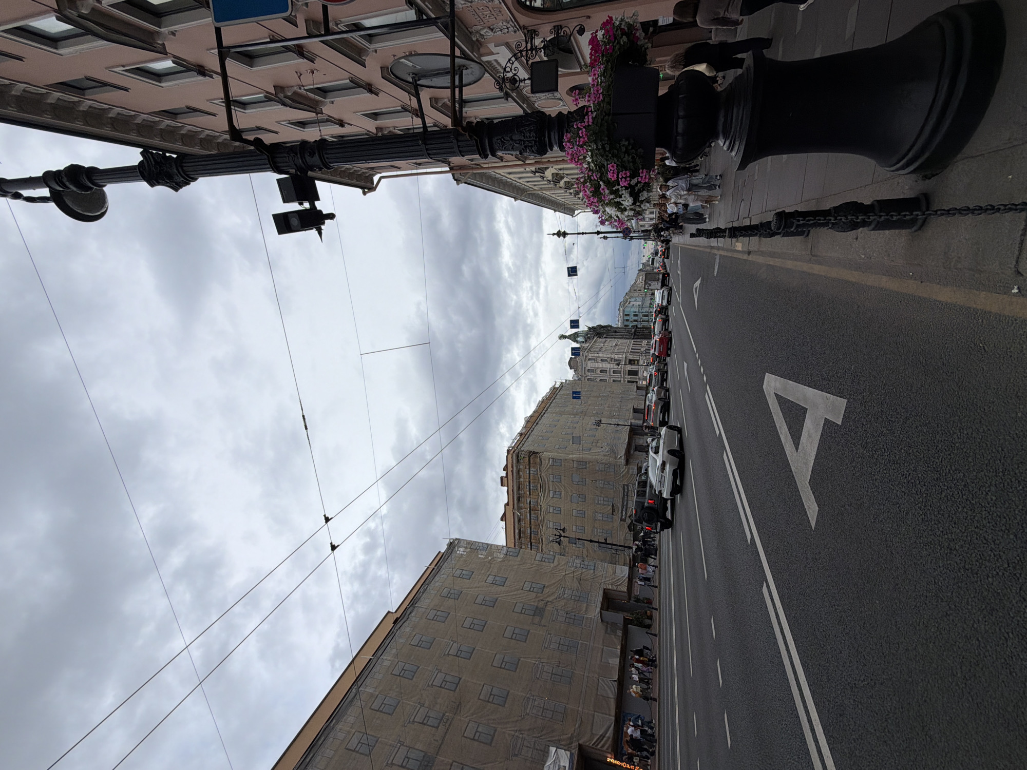 The image depicts a street scene in Saint Petersburg, Russia, likely on Nevsky Prospekt, a major thoroughfare. The sky is overcast with grey clouds. On the left side of the street, two multi-story buildings are covered in scaffolding and beige tarps, suggesting ongoing construction or renovation. The ground floors of these buildings appear to house shops or businesses, with awnings and signs visible. A white road marking in the shape of the letter "A" is prominent on the asphalt road in the foreground.
The road itself has multiple lanes, with several cars driving in the same direction as the camera. Traffic includes a white sedan, a black SUV, and other vehicles. Pedestrians are visible on the sidewalk to the right, walking past shops and cafes. A prominent black lamppost with decorative elements stands on the sidewalk, adorned with hanging planters filled with pink and white flowers. Buildings on the right side of the street are pink and have ornate architectural details. Overhead, a network of electrical wires crisscrosses the sky, indicating the presence of public transportation, likely trolleybuses. The overall impression is of a busy urban environment under a cloudy sky.