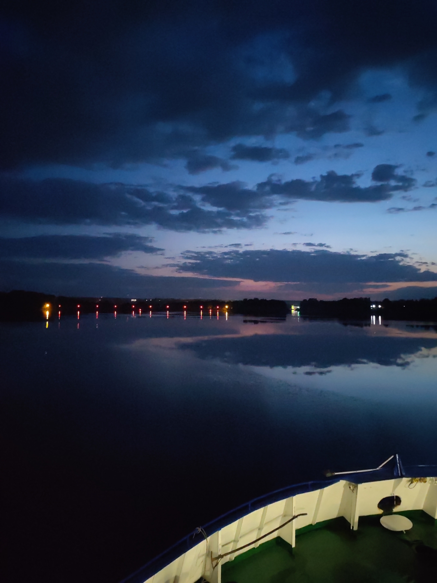 The image depicts a body of water, possibly a river or lake, at dusk or night. The sky is filled with dark, heavy clouds with hints of blue and pink where the sun may have recently set. Lights from a distant shoreline reflect in the water, appearing as rows of red and yellow dots. The foreground shows the bow of a boat or ship, with green flooring and white walls trimmed with blue. The specific location and connection to Ermolino, Russia, cannot be definitively determined without additional information or context.