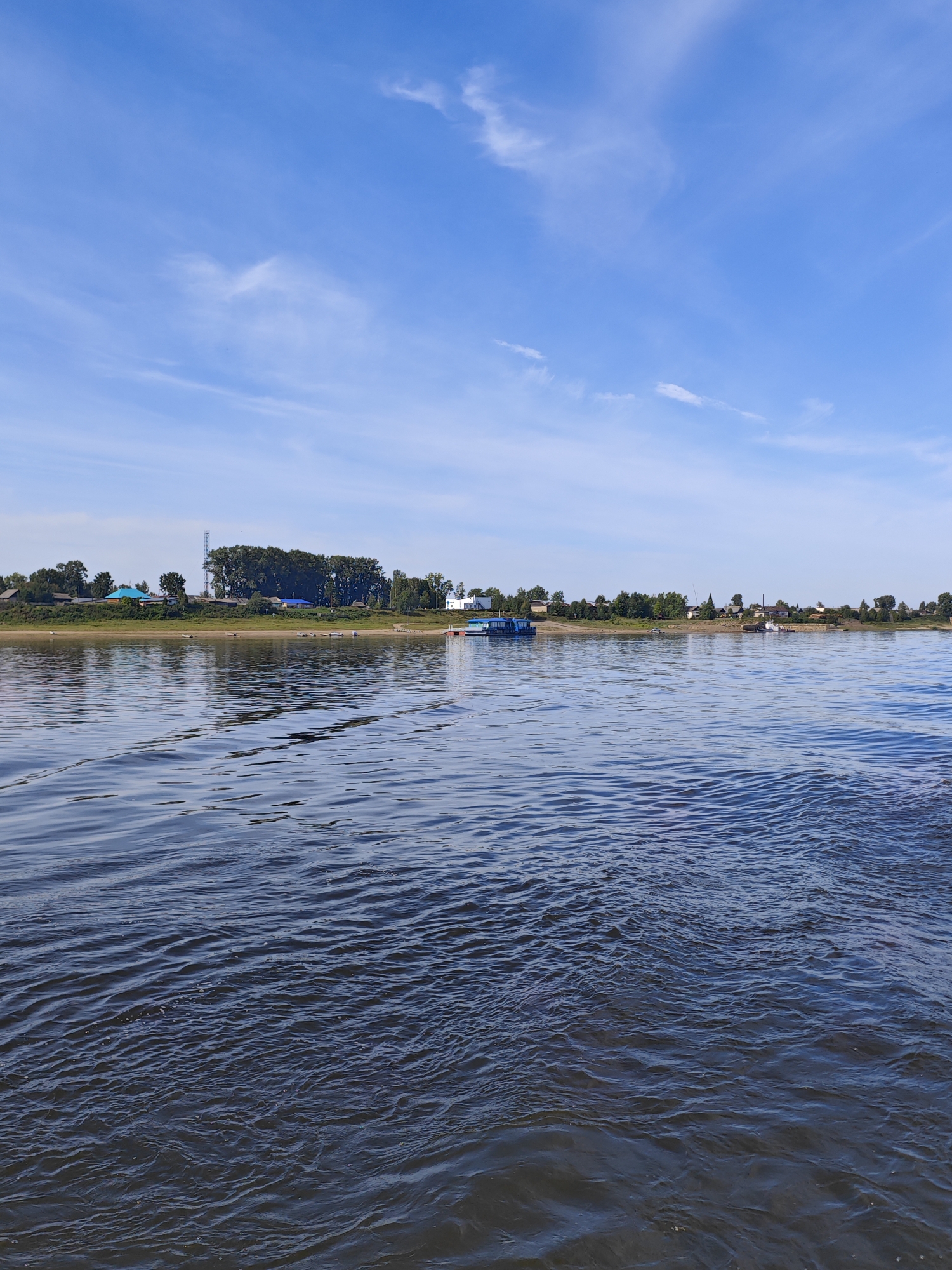 The image captures a wide, calm river with gentle ripples reflecting the clear blue sky and wispy white clouds. On the far bank, a treeline with scattered houses and buildings forms a distant settlement. One building stands out with a bright blue roof. A tall communication tower is visible amongst the trees. Several small boats are moored along the shore, and a larger blue and white vessel is docked further down the river. The water occupies the foreground and midground, with the riverbank and settlement in the background. The scene is tranquil and appears to be a sunny day in a rural or semi-rural setting. It is impossible to confirm the specific location in Yartsevo, Russia, based on the provided image alone.