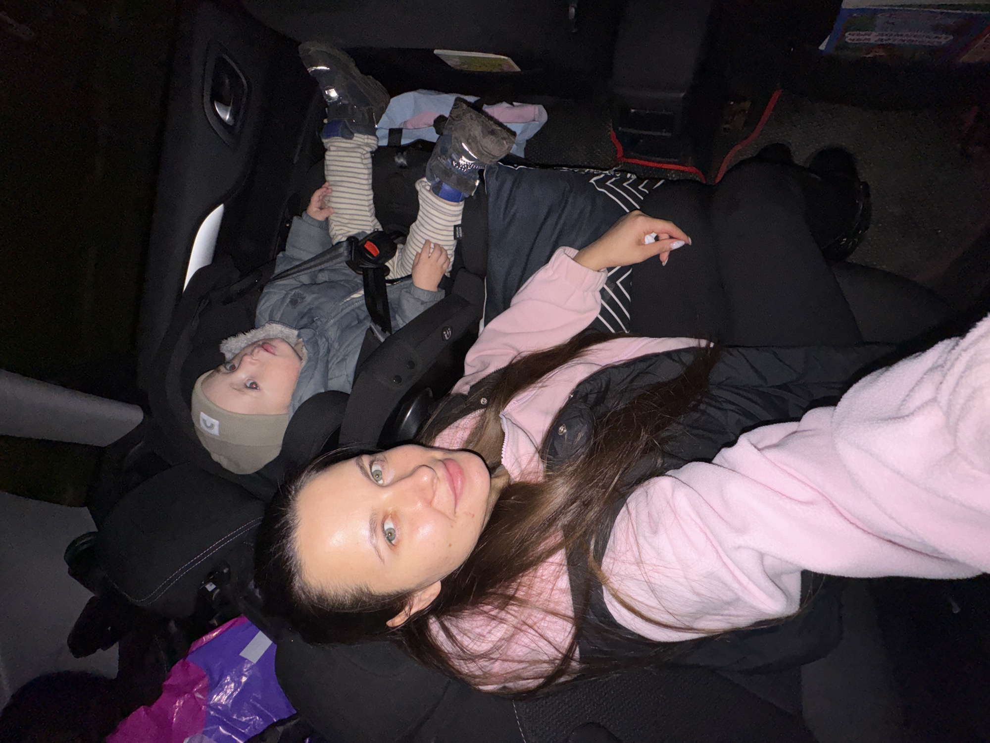 The image shows an adult and a child inside a vehicle. The adult, presumably a woman, is in the foreground, lying on the seat and facing the camera. She has long brown hair and is wearing a pink sweatshirt over a dark jacket. The child is secured in a car seat in the background. The child is wearing striped socks, dark shoes, a gray jacket with fur trim around the hood, and a beige hat. Both are looking towards the camera. There are bags and other items scattered around the vehicle's interior. The location is specified as Красный Маяк, Russia, though there is nothing visible in the image to confirm this. The lighting is dim.
