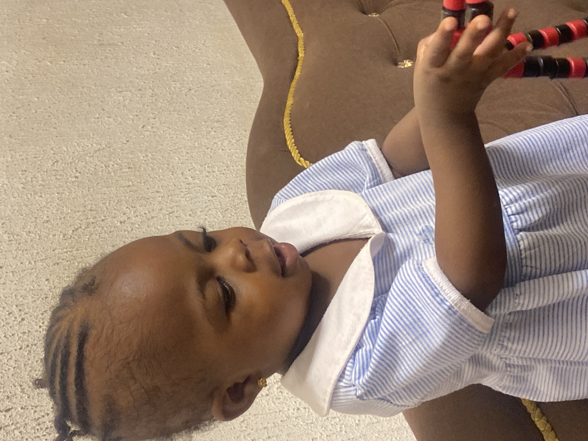 The image shows a young Black child, likely a toddler, with braided hair and wearing a blue and white striped dress. The child is sitting on a brown couch with a gold rope trim, against a textured, light-colored wall. The child is looking down and holding a red and black segmented toy in their hands. The child has a small gold earring in their left ear. The scene appears to be indoors, possibly in a residential setting in Abakaliki, Nigeria.