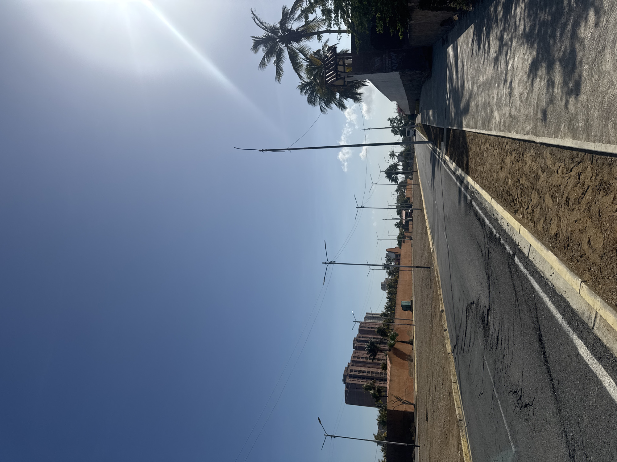 The image shows a street view in Porlamar, Venezuela. The street is paved and has a white line marking the edge of the road. On one side of the street is a sidewalk with tree shadows, and on the other side is an area of dirt and landscaping. Streetlights line the road. In the distance are buildings, including a tall brown building and palm trees. The sky is bright blue with the sun shining.