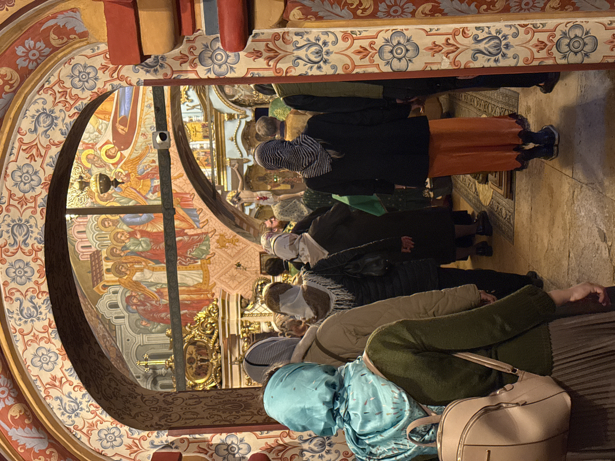 In a rotated interior shot, a group of women is seen praying in a church. The scene is likely in Moscow, Russia due to the style of the church architecture and decoration.
The women are standing in a line, most of them facing forward. They are all wearing head coverings of different colors and patterns. Some are wearing masks. The woman closest to the camera is wearing a blue head covering and a green sweater. There is a beige purse next to her.
The church is highly decorated with paintings and gold accents. The ceiling is arched and painted with religious scenes. There is a security camera mounted on a pillar. The floor is patterned with floral designs.