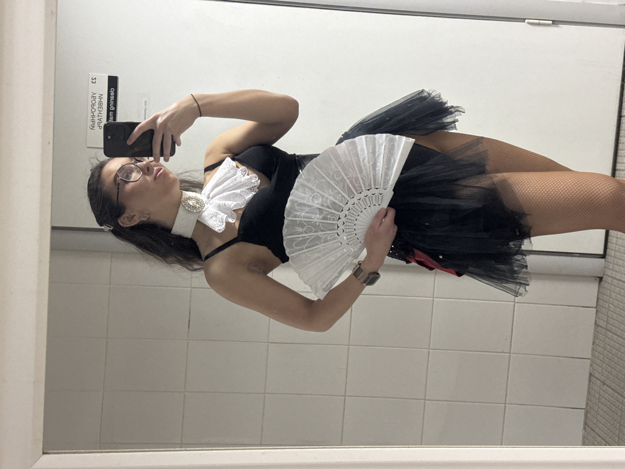 The image shows a woman taking a mirror selfie in a bathroom. She is wearing a black corset-style top with a white ruffled collar, a black tutu skirt, and fishnet stockings. She is holding a white folding fan in her right hand and her phone in her left hand, capturing the reflection. The bathroom appears to have white tiled walls and a grey tiled floor. A sign with Russian text is visible in the mirror's reflection. The location is not specified within the image itself.