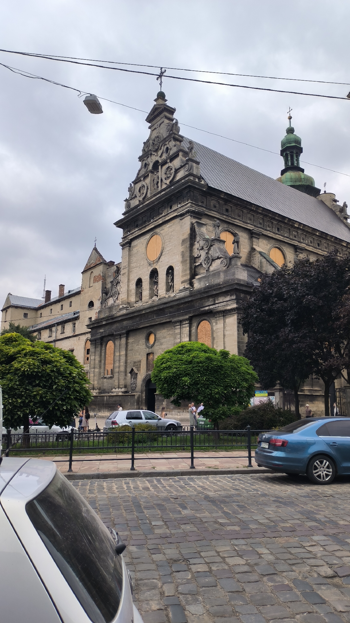This image captures a view of the Church of the Body of God, a prominent architectural landmark in Lviv, Ukraine. The church, constructed from stone, features a grand facade with intricate carvings and decorative elements. Several windows on the facade are boarded up with orange material. A green dome with a cross sits atop one of the church's towers. The foreground of the image is a cobblestone street, with several parked cars. A blue Volkswagen Jetta is visible on the right side of the frame, parked near the sidewalk. A silver hatchback is partially visible on the left. Several trees, one with dark foliage and two with bright green, lush leaves, stand in front of the church. A black metal fence separates the sidewalk from the street. A few people are seen walking and standing on the sidewalk in front of the church. The sky is overcast and gray, suggesting cloudy weather. Overhead, power lines and a street lamp are visible against the sky.