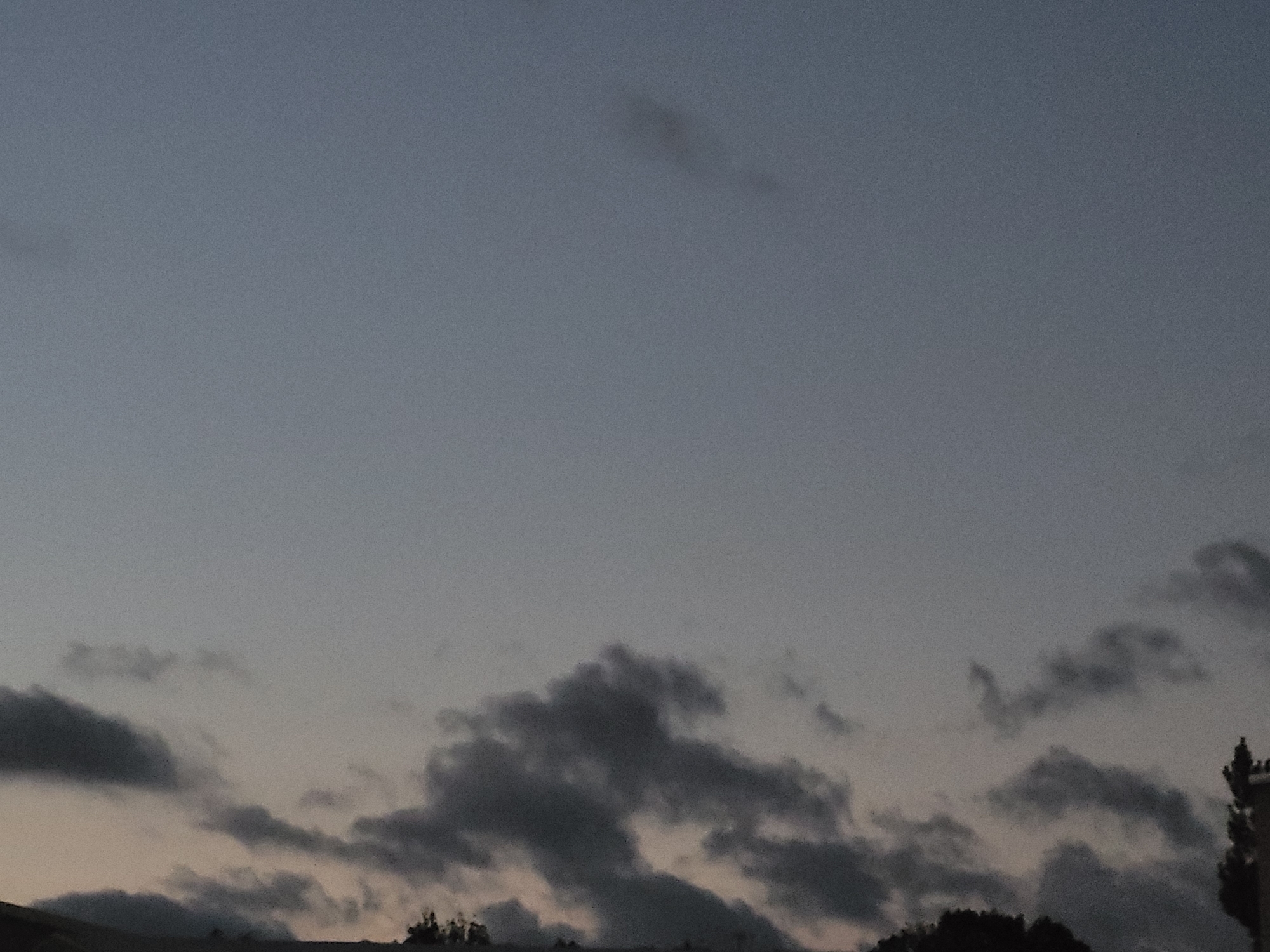 The image depicts a dusky sky with scattered clouds. The sky is a gradient of muted blues and grays, transitioning to a lighter hue near the horizon. Darker, more defined clouds are visible in the lower portion of the frame, obscuring the ground and silhouetting some foliage. A hint of a building or structure is visible on the far right, adding a man-made element to the natural landscape. The overall impression is one of twilight or early evening, with subdued lighting and a somewhat somber atmosphere. There are no people or animals visible in the image. The scene appears to be a natural landscape with some urban elements present. There is no specific indication of the city of Ozolnieki, Latvia, within the image itself.