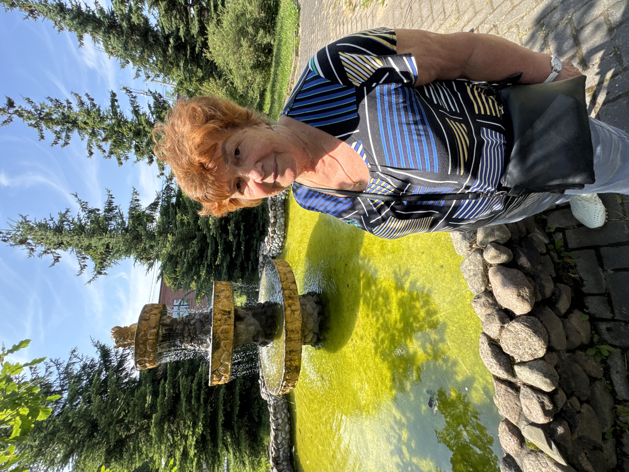 The image shows an elderly woman with reddish-blonde hair standing outdoors next to a fountain. She is wearing a black, blue, yellow, and white striped shirt and blue jeans. A black purse is slung over her shoulder. Her expression appears neutral. The fountain has multiple tiers and is surrounded by decorative stones. The water in the fountain is a vibrant green, suggesting algae growth. In the background, there are evergreen trees and a blue sky with wispy clouds. The scene suggests a sunny day, likely in the late morning or early afternoon, given the lighting. The paved walkway behind the woman indicates a public area or garden. The location context provided is Lomonosov, Russia, which is consistent with the presence of evergreen trees and the style of the fountain and walkway. There is no visible text in the image.