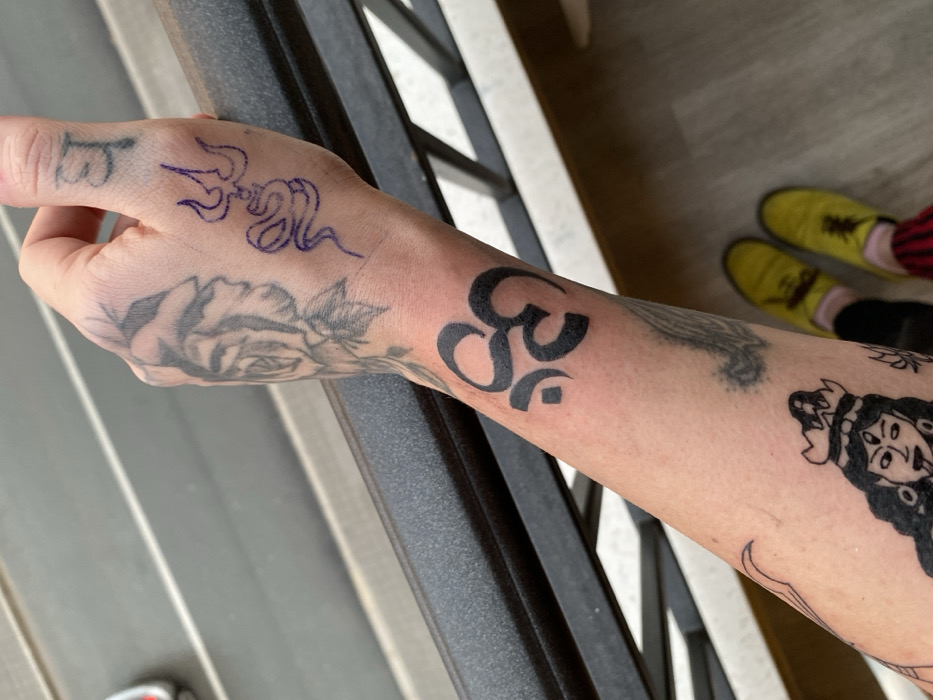 A fair-skinned arm and hand feature multiple tattoos. On the wrist is a solid black Om symbol. Further down the arm is a black ink tattoo of a face. The hand shows a rose in faded black ink, a trident entwined with a snake in blue ink, and letters on the thumb. Yellow shoes and striped socks appear at the top of the image. The background shows a balcony railing and a gray floor.